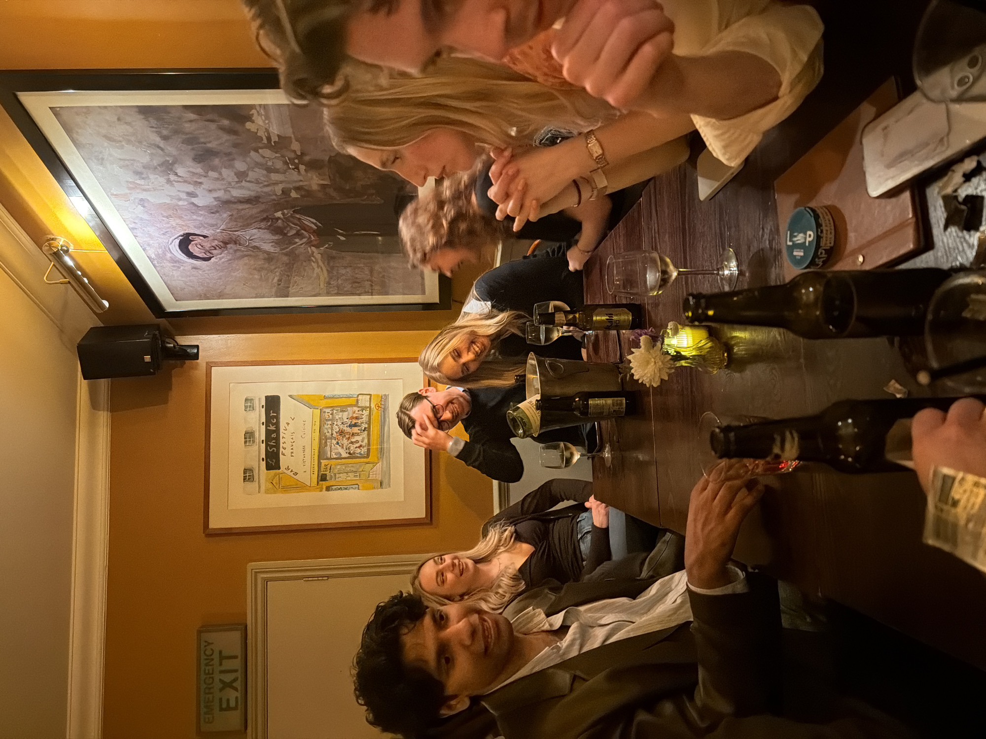 A group of about 7-8 people are seated around a dark wooden table in what appears to be an upscale establishment. The scene is illuminated by warm ambient lighting. Several wine bottles and glasses, along with a small vase of flowers, are scattered across the table. The individuals are engaged in conversation and appear to be enjoying a social gathering, with some looking directly at the camera. The establishment features artwork on the walls, including a large framed painting and a smaller framed print displaying French text, possibly indicating a French restaurant or bar. An "Emergency Exit" sign is visible near a door. The room has a warm, cozy ambiance. Due to the presence of the print on the wall with the text "L Shaker Festival Français", it is possible that this photograph was taken at L Shaker, a restaurant located in London, UK.