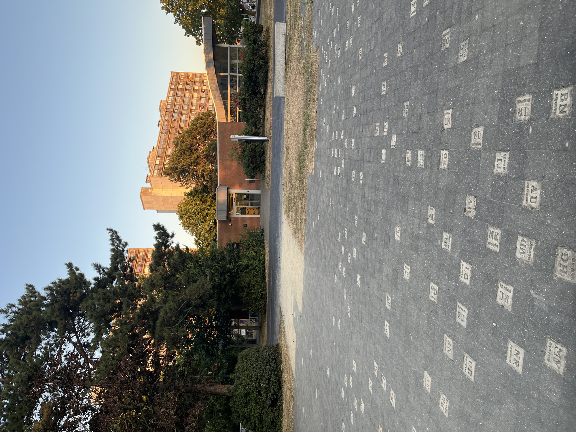 The image shows a view of the street pavement in Ostend, Belgium, where each paver stone is imprinted with a two-letter code and a four-digit number. The pavement is laid out in a grid pattern, and the pavers are a dark gray color. In the background, there is a brick building with glass windows and a covered walkway. To the left of the building, there are trees and bushes. Further in the background, there is a tall, multi-story building. The sky is clear and blue, indicating a sunny day. The scene appears to be an outdoor public space.