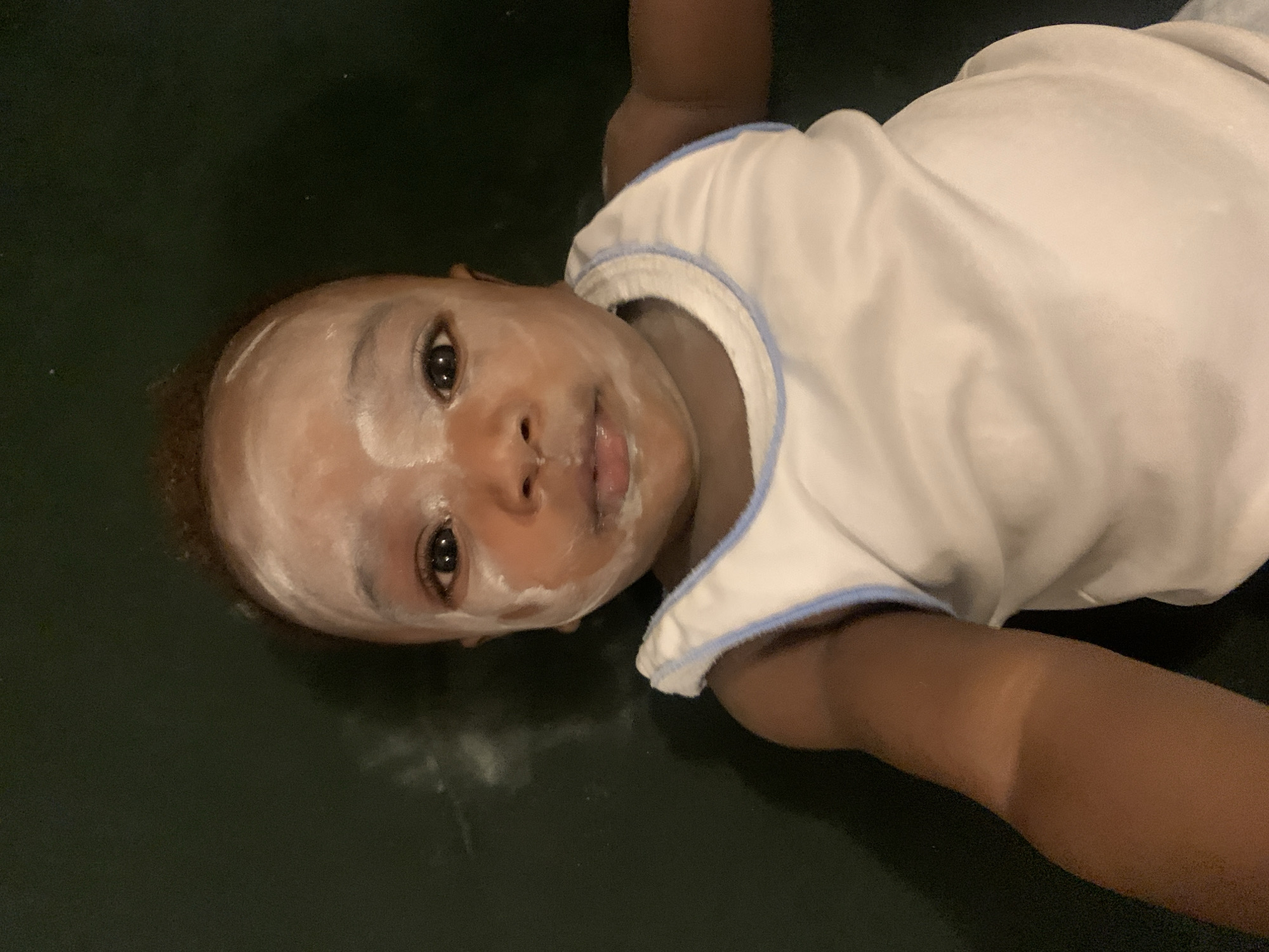 A baby lies on a dark, possibly green surface. The baby has a white substance on its face. The baby is wearing a white shirt with light blue trim. The location is not discernible from the image.