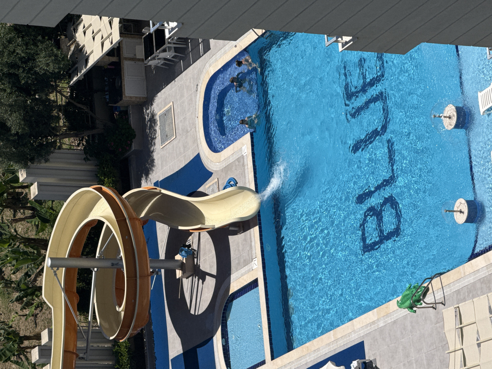 This is an aerial, slightly oblique view of a resort or hotel swimming pool complex, brightly lit by sunlight. The scene is dominated by a large, blue rectangular swimming pool on the right side of the image, featuring the word "BLUE" written in dark letters on its bottom. A smaller, irregularly shaped pool is connected to the main pool by a grey tiled patio area.
At least three people are visible in the smaller pool, and one person is captured in motion, emerging from the end of a large, spiraling orange and beige water slide into the main pool, creating a splash. The water slide descends from the left side of the frame. Nearby, blue hosing is coiled on the ground.
The pool deck is paved with grey tiles. A green frog statue is positioned on the edge of the main pool, and two decorative circular water features are visible within the pool itself. In the background, to the left, are buildings with balconies or awnings, partially obscured by green trees and foliage. Several white lounge chairs are scattered around the pool area. Strong shadows cast by the structures and slide indicate a clear, sunny day, suggesting a leisurely and active holiday environment.
Location context: Unknown, Unknown