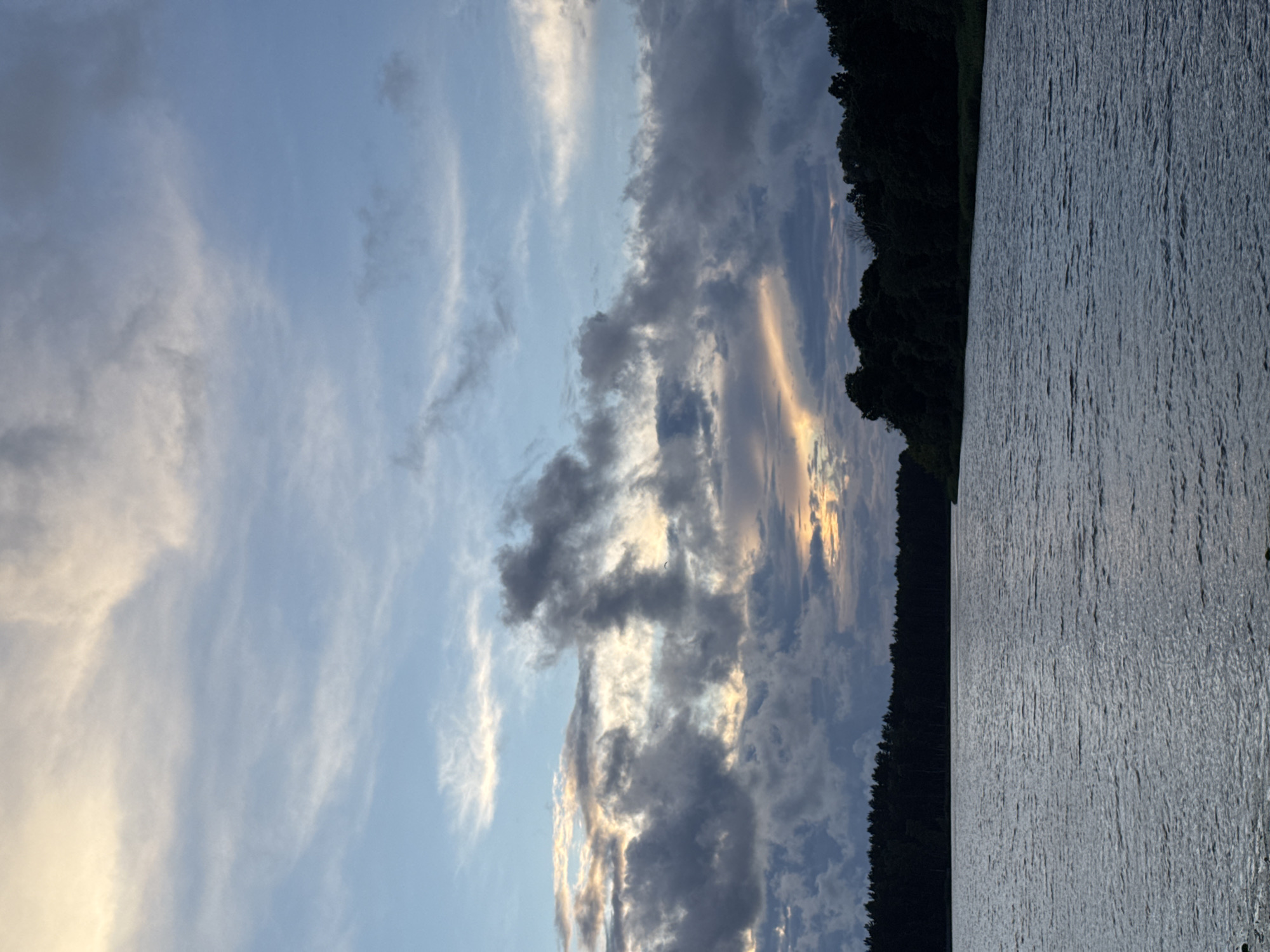 A wide shot captures a tranquil lake in Carnikava, Latvia, at dusk. The water's surface is slightly rippled, reflecting the muted colors of the sky. A dense, dark line of trees forms the far bank of the lake, with the forest appearing silhouetted against the horizon. Above the trees, the sky is a canvas of dramatic clouds. Lighter, wispy clouds are interspersed with darker, heavier formations, creating a sense of depth and movement. A soft glow of sunset light peeks through a gap in the clouds, casting warm hues of orange and yellow onto the sky. There are no people or man-made structures visible in this natural landscape.