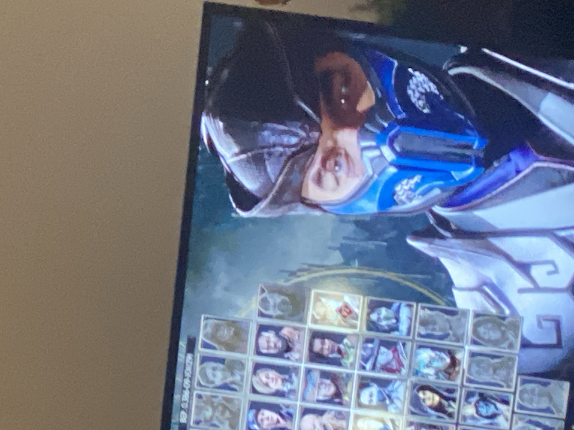 The media file displays a close-up, rotated view of a video game character selection screen. The primary subject is a character with a dark helmet, blue and white armored clothing, and a visible face with a reflected gleam in their eye. Below the character, a grid of smaller selectable character portraits is visible. These portraits are mostly blurred but appear to depict various individuals, some in armor and some with different hairstyles and facial features.
The overall scene is a digital interface, likely from a fighting game, given the character portrait grid. The lighting appears to be artificial, coming from the screen itself, which casts a soft glow on the character and the surrounding interface. There are no external environmental cues like weather or time of day. The mood conveyed is one of anticipation, common in game selection menus.
Visible text includes a string of alphanumeric characters and symbols along the left edge of the screen, possibly a file name or code: "SEP.0.356-09-104759". The character portraits themselves are too small and blurred to decipher any text within them. The background of the main character is dark and indistinct, with some subtle lighting effects. The image is tilted, making a direct interpretation of orientation difficult.
