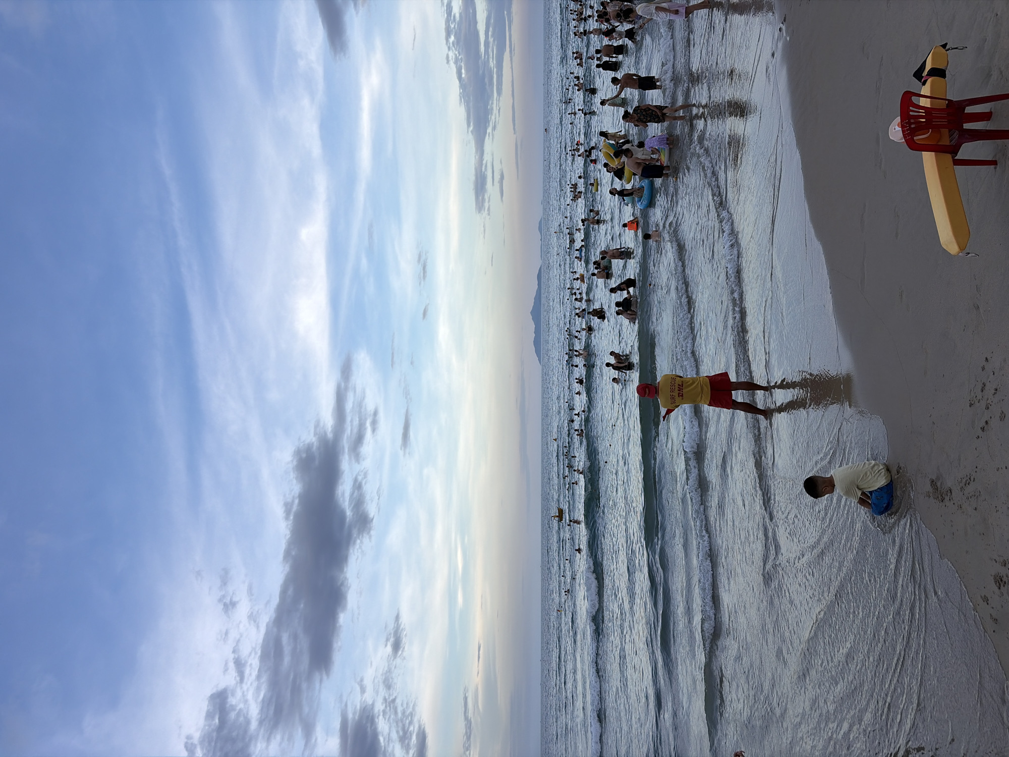 The image depicts a crowded beach scene in Đà Nẵng, Vietnam, during what appears to be late afternoon or early evening, given the soft lighting and the colors of the sky. The ocean is active with moderate waves, and numerous people are swimming, wading, and playing in the water. In the foreground, a young boy is sitting on the wet sand near the water's edge, facing away from the camera. To the right, a lifeguard in a yellow shirt with "SURF RESCUE" and "DHL" branding, and red shorts, stands facing the ocean, one arm raised as if signaling. In the far distance, a faint outline of mountains is visible on the horizon. Scattered throughout the water are inflatable toys and some people are on them. On the beach in the lower right corner, a yellow rescue float and a red plastic chair are positioned. The sky is a blend of blue with scattered clouds, some of which are dark and wispy, suggesting the potential for changing weather. The overall atmosphere is one of a busy recreational day at the beach.
