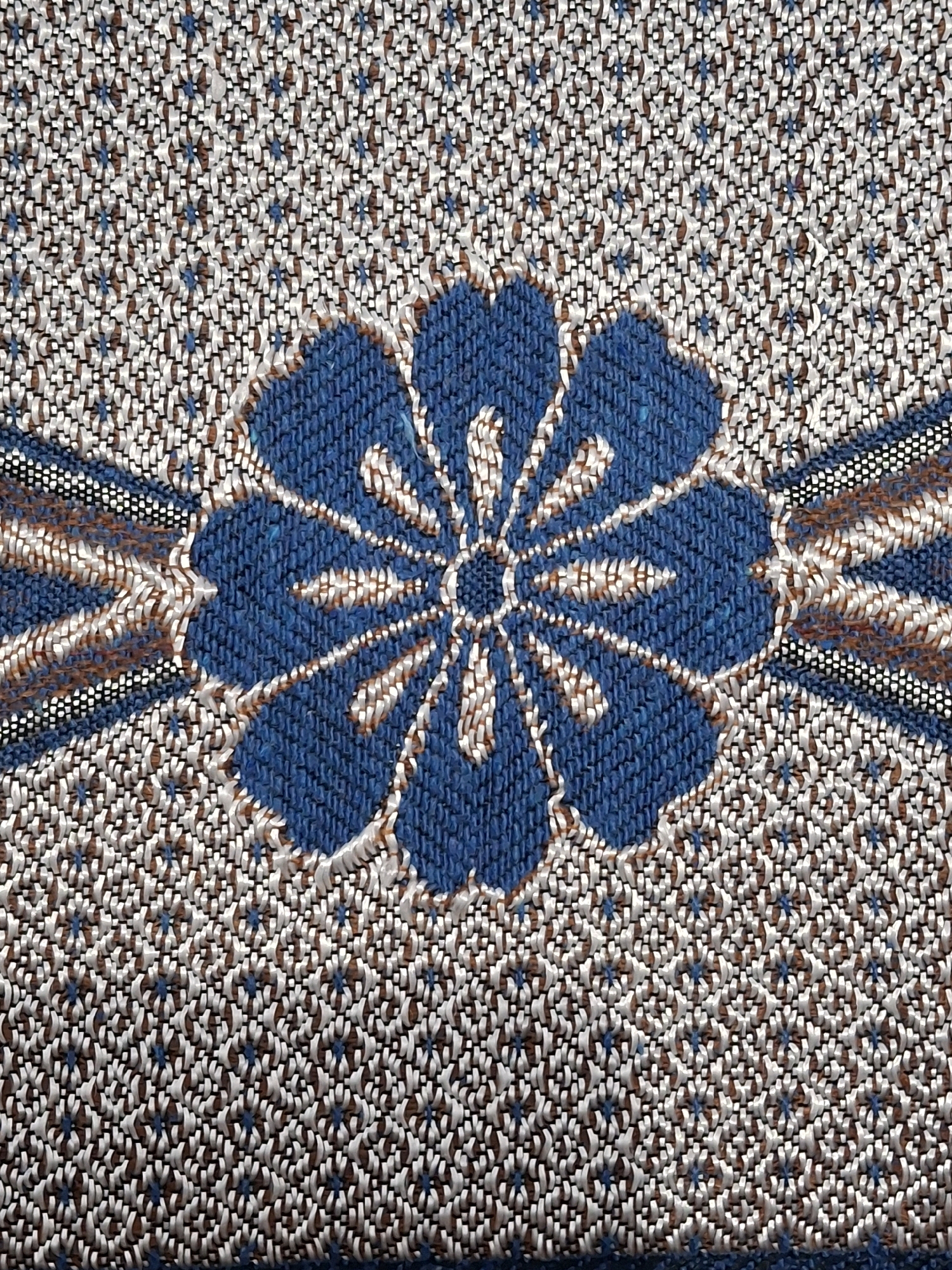 The image is a close-up, overhead view of a woven fabric with a floral motif. The central element is a large, stylized blue flower with eight petals, each outlined in white and tan threads. Extending from the sides of the flower are woven bands of black, white, and tan threads that create a symmetrical pattern. The background fabric is densely woven with a repeating pattern of small, intricate shapes formed by white, tan, and small blue threads. The texture of the fabric is prominent, showing the raised threads and the weave structure. The lighting appears to be even, highlighting the details and colors of the textile. There are no people, animals, or environmental elements visible in the image; the focus is solely on the fabric's design and texture. The image does not contain any elements that can be used to determine its location within Lumajang, Indonesia, or any other geographical area.