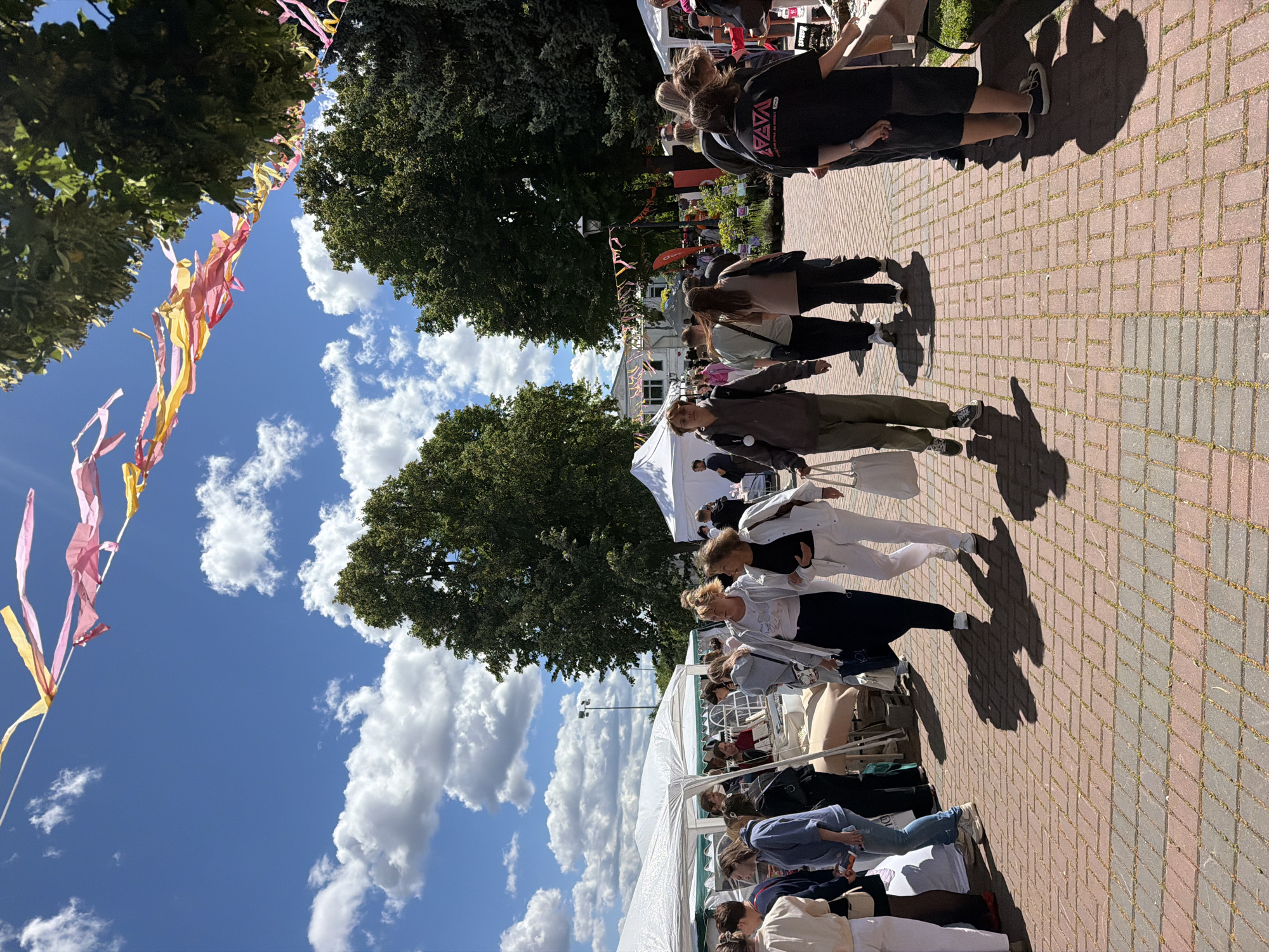 The image depicts an outdoor event, possibly a fair or market, with several people browsing stalls. The scene is set on a brick pathway lined with trees. Colorful streamers are strung across the pathway, adding a festive atmosphere. The weather appears to be sunny with scattered clouds, suggesting it's daytime. Several white tents or market stalls are visible, with merchandise displayed on tables. People are seen walking, standing, and interacting, suggesting a relaxed and casual event. Some individuals are looking at items on display, while others are conversing. The overall mood seems positive and engaging. In the background, a building with a white facade is visible, possibly indicating a public space or cultural venue. The image has a slightly tilted perspective, making it appear as if the viewer is looking up from the ground. No specific text is clearly legible in the image, though some logos or signs might be present on the stalls or clothing. The setting is consistent with a warm, fair-weather day in a town or city park.