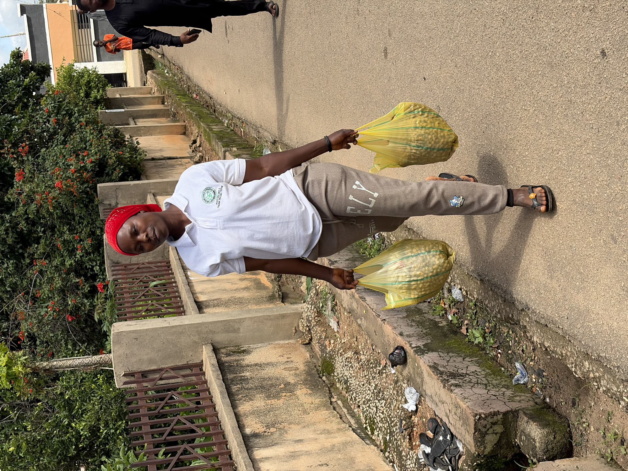 A medium shot shows a young woman walking on a paved road in Jos, Nigeria, carrying two yellow plastic bags filled with food. She wears a red headscarf, a white polo shirt with a logo, and grey sweatpants. Her feet are clad in black flip-flops with yellow flower accents. The woman walks towards the viewer, looking to her left. To her right, a ditch runs along the side of the road, containing scattered debris and overgrown weeds. A brown metal fence with a concrete wall separates the road from a dense green bush with red flowers. In the background, two men are visible walking away from the camera; one wears a black garment and the other an orange shirt. The overall scene depicts an ordinary day in a Nigerian city, with the woman engaged in the task of carrying her purchases.