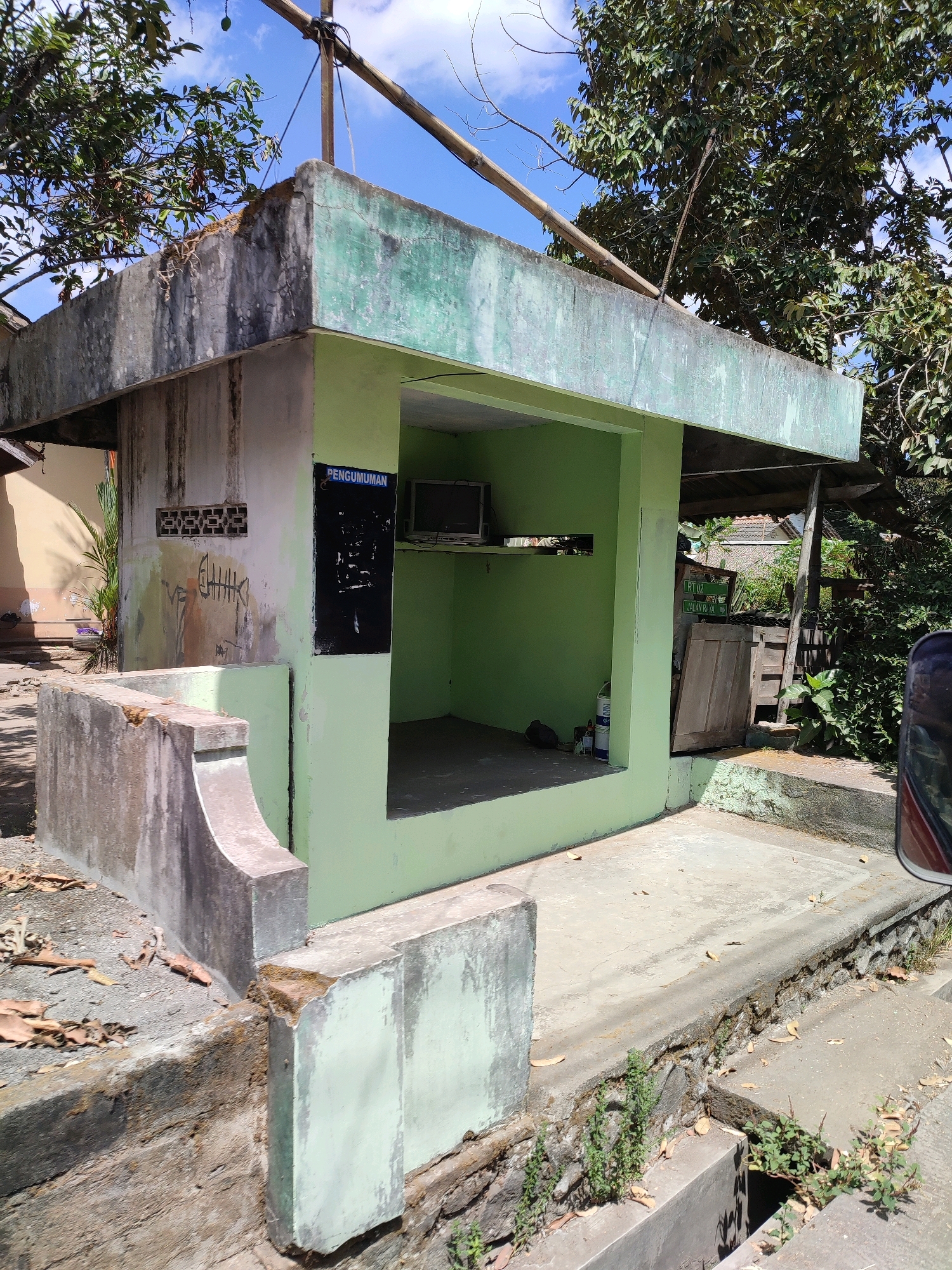 The image depicts an abandoned small concrete structure, likely a former kiosk or information booth, situated in a semi-urban or rural environment. The structure is painted a faded light green and shows signs of disrepair, with peeling paint and cracks in the concrete. A dark poster with the word "PENGUMUMAN" (Announcement) is affixed to one wall, with illegible scribbles beneath it. Inside the open-fronted structure, a small, old television set is visible on a shelf, along with some containers.
The ground in front of the structure is paved with concrete, which also appears weathered and cracked. To the left, a low concrete wall or barrier, also showing wear, runs along the edge of the paved area. Vegetation, including dry leaves and small plants, is scattered on the ground and growing in cracks.
In the background, a large tree with dense foliage dominates the upper right portion of the image, with its branches extending over the structure. Other elements visible in the background include a glimpse of a building with a tiled roof, wooden planks, and a street sign indicating "RT 02 JALAN KYA". A portion of a red vehicle is visible on the far right edge of the frame, suggesting it is parked nearby. The sky above is bright blue with scattered white clouds, indicating a sunny day. The overall impression is one of neglect and abandonment in a tropical setting.