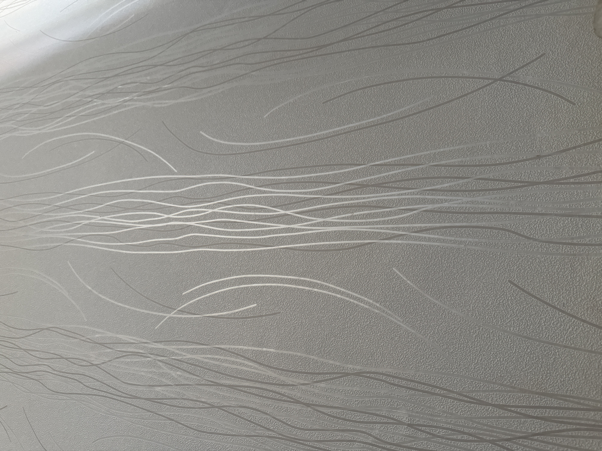 The image shows a close-up of textured wallpaper with a pattern of thin, vertical, wavy lines. The wallpaper is a light gray color. The texture appears to be a raised, stippled effect. The wavy lines are a slightly lighter shade of gray or silver and are subtly raised or have a sheen that catches the light, creating a subtle contrast with the background. The lighting suggests a light source from the top right, casting a slight gradient across the surface and highlighting the texture and lines. There are no people, animals, or specific objects visible other than the wallpaper itself. The location in Anadyr, Russia, is not discernible from the image, as it depicts a generic interior surface.
