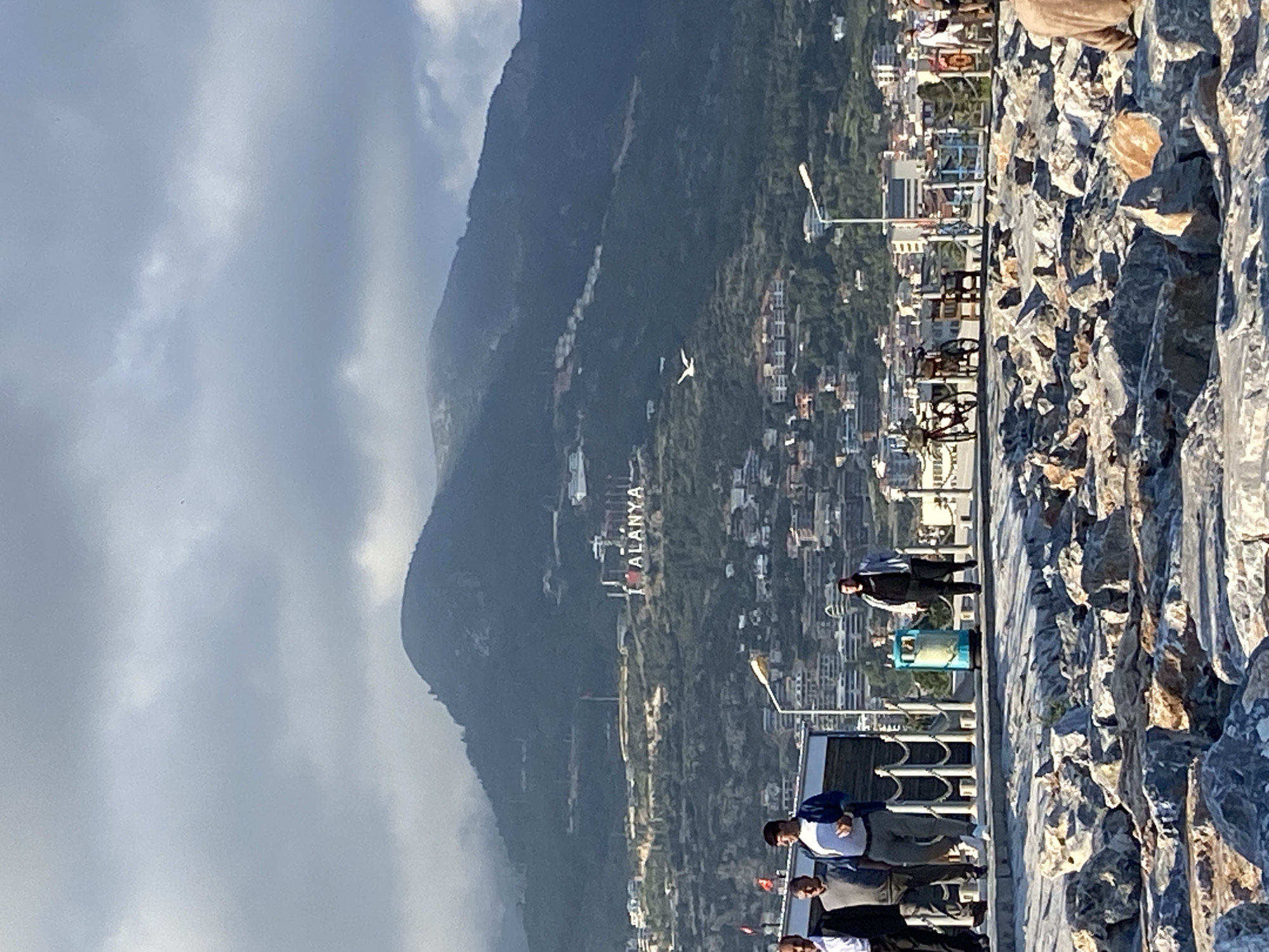 This vertical image captures a daytime scene in Alanya, Turkey, featuring a mountain, a city built into its slopes, and a waterfront promenade. The foreground consists of a rocky embankment made of large, light-colored stones. Adjacent to this is a paved promenade with a white railing and streetlights.
Several people are visible on the promenade. A group of men, some wearing casual attire like shirts and vests, stand near the railing, seemingly observing the view. One individual in a dark jacket is seen further down the promenade. A blue container or kiosk is situated on the walkway, and a bicycle leans against the railing in the distance.
In the mid-ground and background, a large, lush green mountain rises, its slopes dotted with numerous buildings that make up the city of Alanya. Prominently displayed on the mountain face are large white letters spelling out "ALANYA". A white bird, possibly a seagull, is captured in flight against the mountain's backdrop. The sky above is partly cloudy, with patches of brighter areas indicating sunshine. The scene suggests a clear and pleasant day in the coastal city.