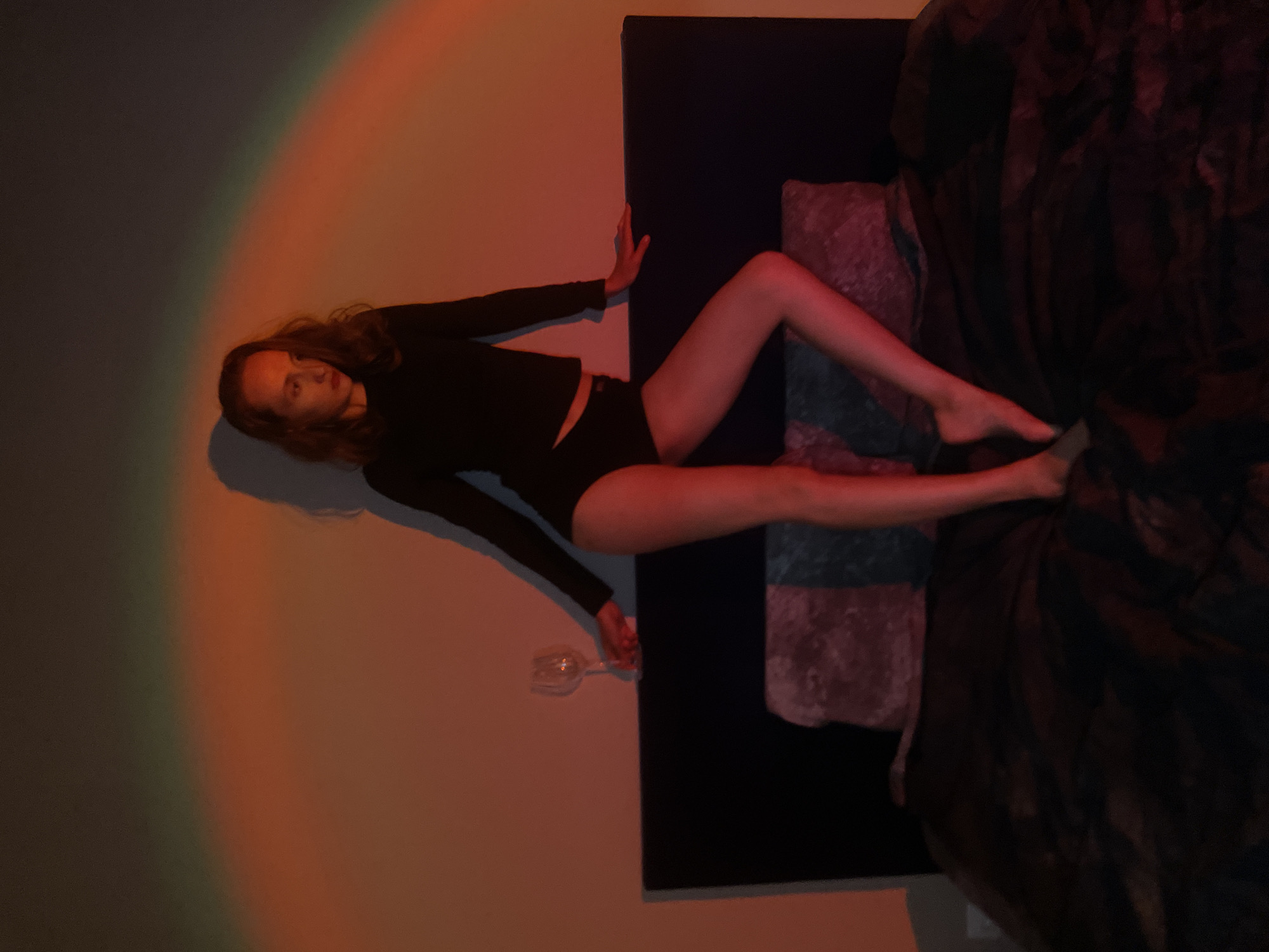 A woman lies on a bed next to a wall in a room with a rainbow light. She is wearing a black long-sleeved shirt and black underwear. She holds a wine glass in her left hand. The bed has a dark headboard and a dark comforter. The location is not specified.
