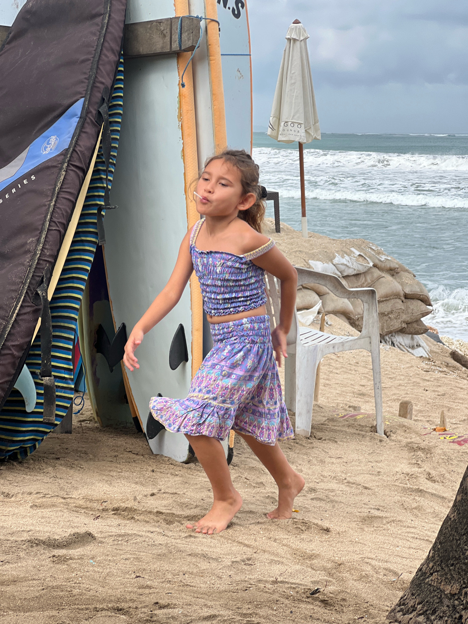A young girl with dark hair is standing on a sandy beach, her eyes closed and a lollipop in her mouth, as if she is blowing a bubble. She is wearing a purple and white patterned off-the-shoulder top and a matching skirt. She is barefoot and her body is angled to the right, with her arms slightly outstretched. Behind her, several surfboards are propped up, leaning against a light blue wall. One surfboard bag is black with a blue and grey logo. A white patio chair is visible behind her, and beyond that, a pile of sandbags secured by netting. Further in the background, the ocean is visible with moderate waves under an overcast sky, suggesting it is either early morning or late afternoon. An umbrella stands on the beach to the right of the chair. A sign on the umbrella reads "GOO LOUNGE." The scene is Kuta, Indonesia.
