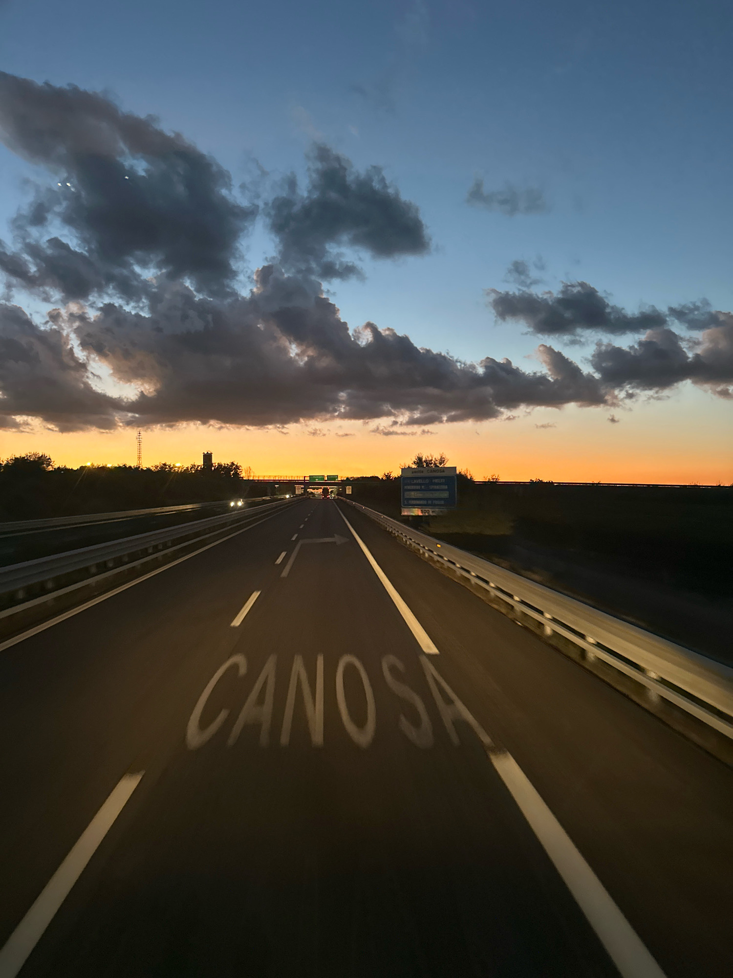 This is a nighttime photograph taken from a moving vehicle on a highway, likely approaching or within the vicinity of Canosa di Puglia, Italy, given the painted text on the road. The sky displays a gradient of deep blue at the top transitioning to a vibrant orange and yellow sunset glow near the horizon. Dark, dramatic clouds are scattered across the sky, catching the last of the daylight. The highway stretches forward, illuminated by artificial lights and the fading sunset. White lane markings are clearly visible on the asphalt. Prominently painted in white on the road surface are the letters "CANOSA," indicating a destination or area. On the right side of the highway, a guardrail runs parallel to the road. In the distance, road signs and streetlights are visible, along with the silhouettes of trees and structures against the colorful sky. A few vehicles with their headlights on can be seen in the distance, traveling in the same direction. The overall atmosphere is one of travel and approach to a destination at dusk.