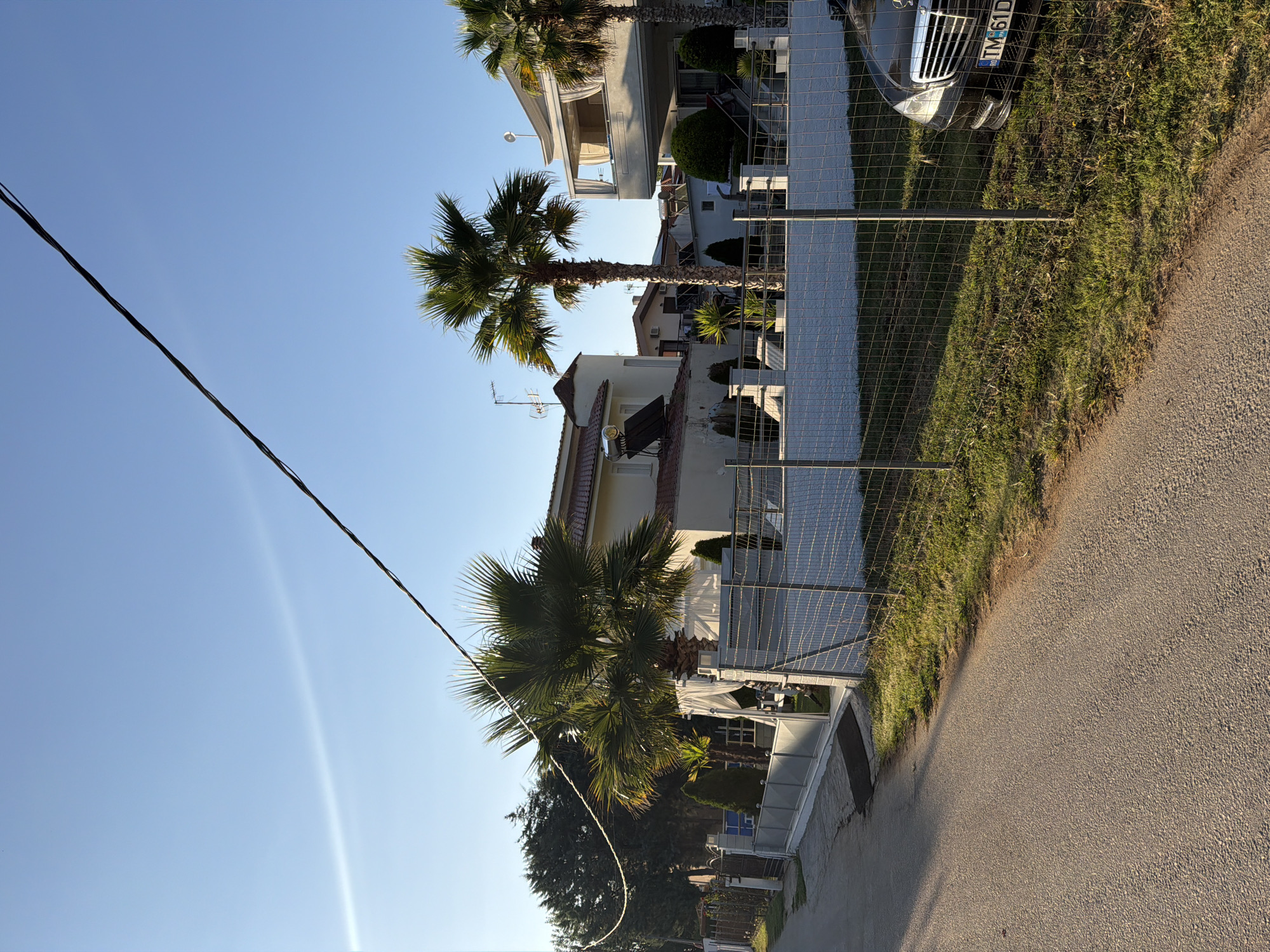 The image shows a street scene in Golden Beach, Greece, during daylight under a clear blue sky. A paved road runs along the left side of the frame, bordered by a grassy verge and a wire mesh fence. Beyond the fence, several modern buildings with white exteriors and red tile roofs are visible, interspersed with palm trees and manicured greenery. One building features a rounded tower with a solar water heater on its roof. Another building has a second-story balcony with white curtains. A black Mercedes-Benz sedan with a license plate starting with "TM 61" is parked on the right side of the frame, partially visible behind the fence. Power lines traverse the upper portion of the image. The scene appears to be in a residential or resort area.