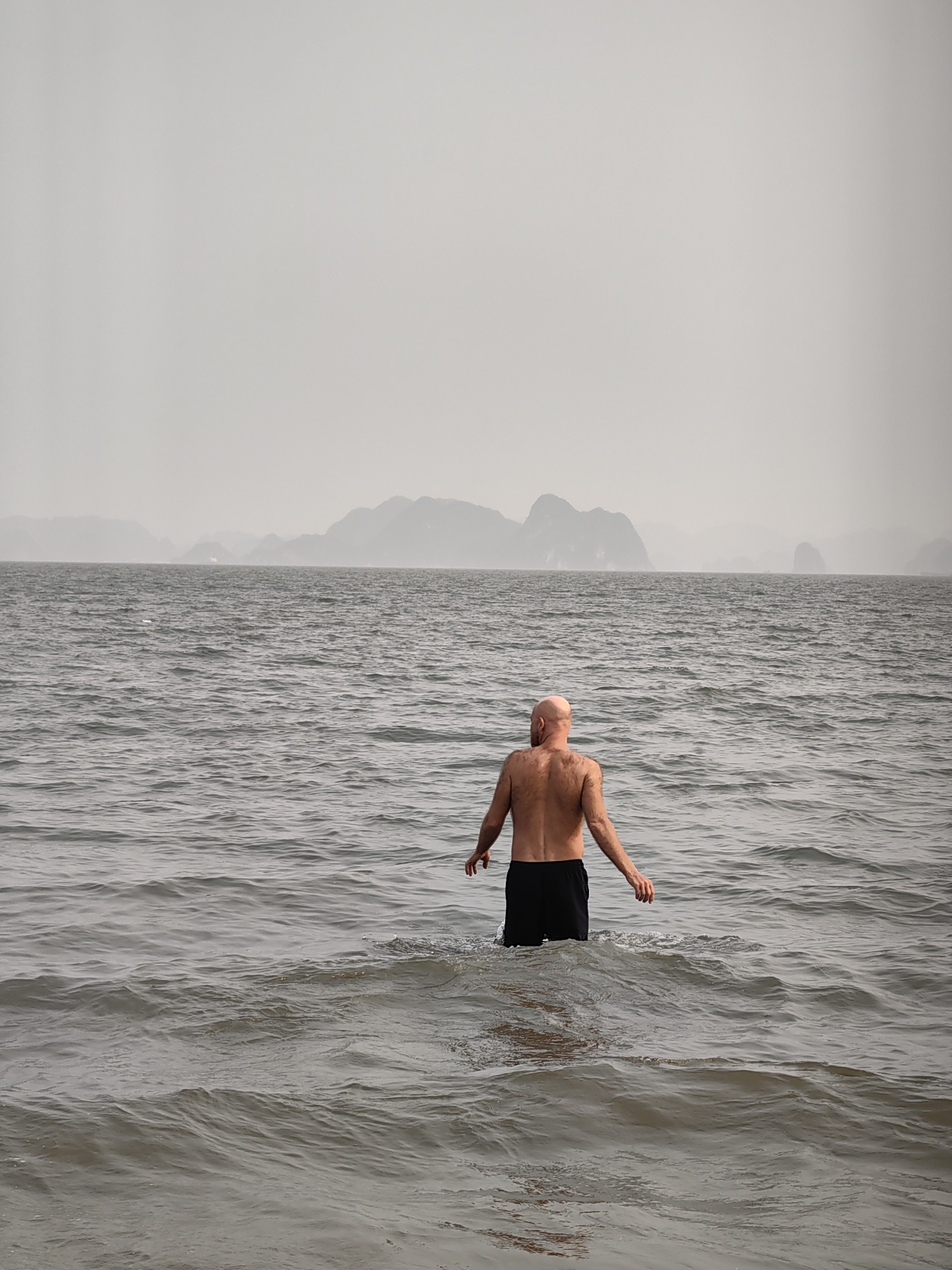 A bald man stands in the sea up to his waist. He is wearing black shorts. The sea is choppy with small waves. In the distance, islands rise out of the water in the hazy fog. The location is in the city of Bai Chay Ward, Vietnam.
