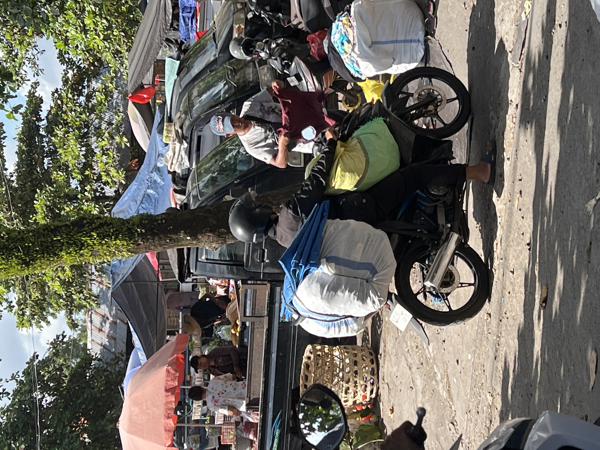 Captured in the vicinity of Singakerta, Indonesia, the photograph reveals a bustling street scene. Two motorbikes heavily laden with bags are parked in front of a row of trucks. Individuals are present near the motorbikes. One person is seated on a motorbike adjusting clothes. Others are present near the storefronts. Trees frame the street. The lighting suggests a sunny day.