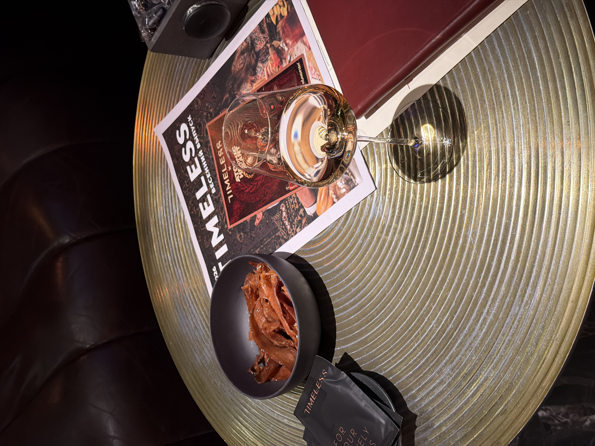 A high-angle shot captures a well-lit scene on a golden circular table. A wine glass filled with amber-colored liquid sits atop a magazine with the word "TIMELESS" prominently displayed. Adjacent to the magazine, a red book rests on the table.
In the foreground, a dark bowl contains what appears to be strips of dried meat or jerky. A stack of black packets, bearing the "TIMELESS" logo, are also present on the table. The table itself has a ridged texture. A dark, textured surface is visible in the background, possibly a leather couch or chair.
The location is Unknown, Unknown.