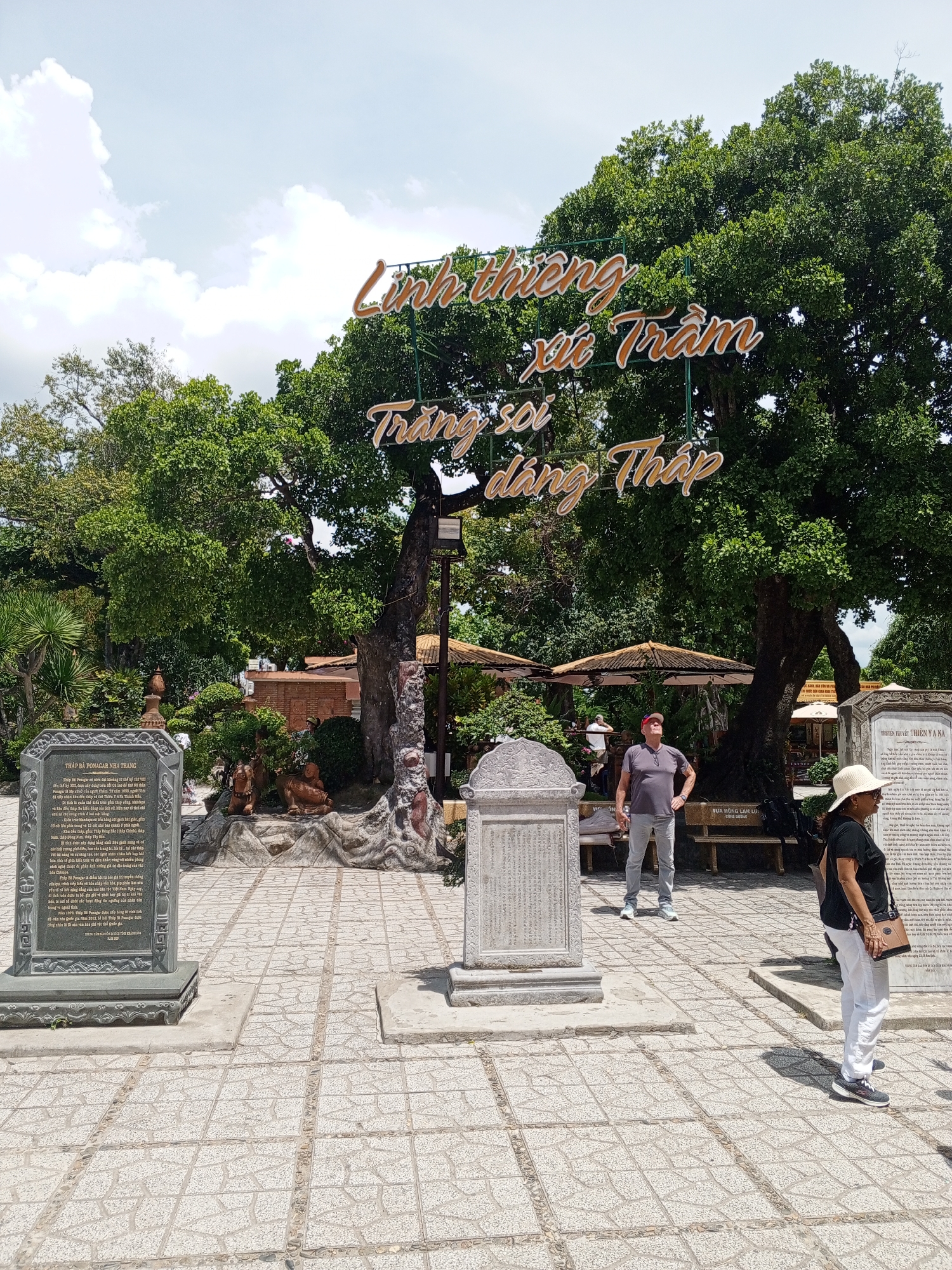 In the city of Nha Trang, Vietnam, within Bac Nha Trang Ward, this outdoor scene showcases a pathway paved with light-colored square tiles, interspersed with decorative stone borders. A large, mature tree with dense green foliage dominates the background, its branches partially obscured by a prominent sign with gold lettering that reads "Linh thiêng xứ Trầm”. Below this, another sign reads "Trang soi dâng Tháp”.
In the foreground, to the left, stands a grey stone monument inscribed with text in Vietnamese. To its right, a smaller, similarly styled monument is visible. Two individuals are present: a man wearing a red cap, grey t-shirt, and grey pants stands in the center, looking upwards, with his hands on his hips. To his right, a woman wearing a white hat, dark t-shirt, and white pants stands facing right, a brown bag slung over her shoulder. Behind them, a wooden bench is situated.
Further into the background, beyond the main tree, are more trees, some covered seating areas with thatched roofs, and brick structures. The sky is bright and appears partly cloudy. The overall impression is of a well-maintained historical or cultural site under a clear, sunny sky.
