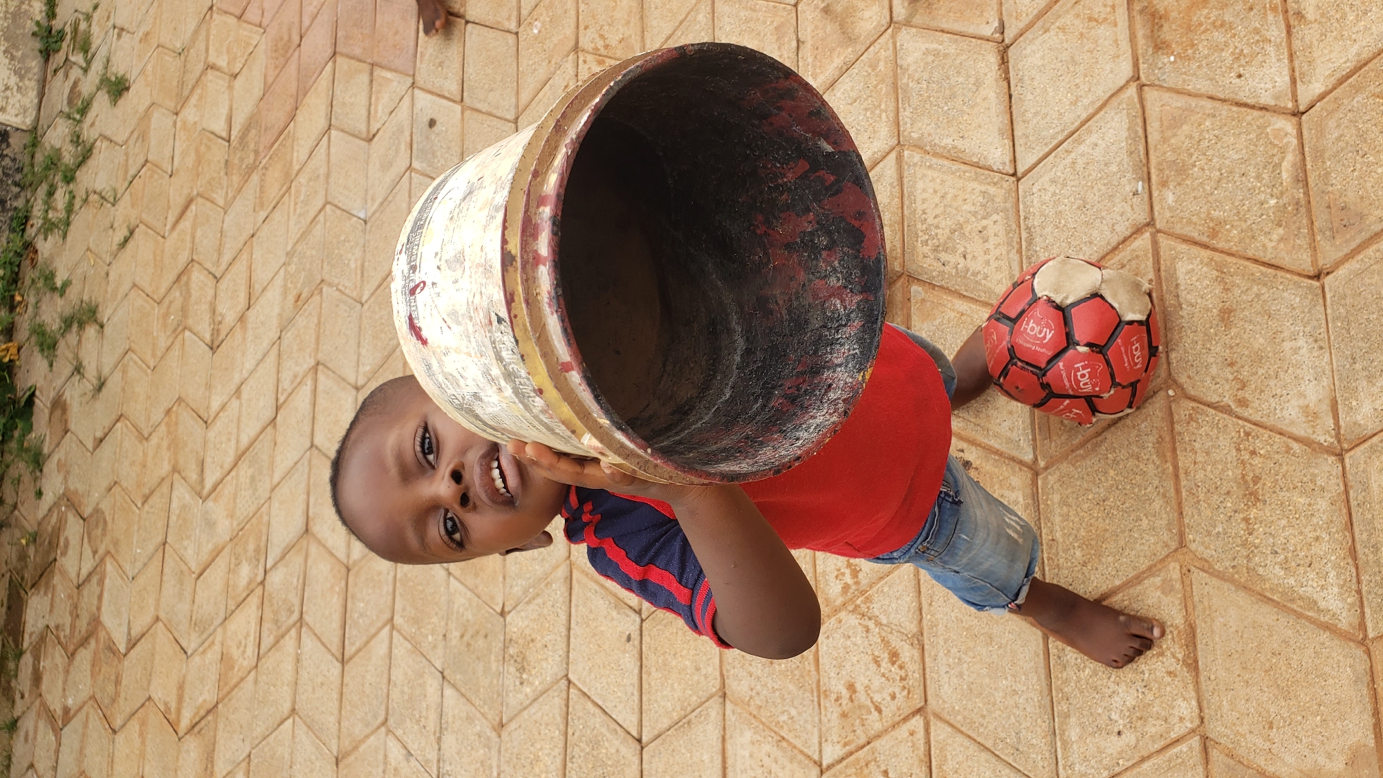 A young boy stands on a diamond-patterned concrete patio, holding a large, weathered cylindrical container by the rim. He is looking directly at the camera with an open mouth, as if speaking. The boy wears a red t-shirt with blue and red striped sleeves and light blue denim shorts. To his right, a red and white soccer ball rests on the patio. The background consists of the patterned patio and some sparse green vegetation. The scene is outdoors, likely in a residential area, given the patio and the presence of a child with a toy.