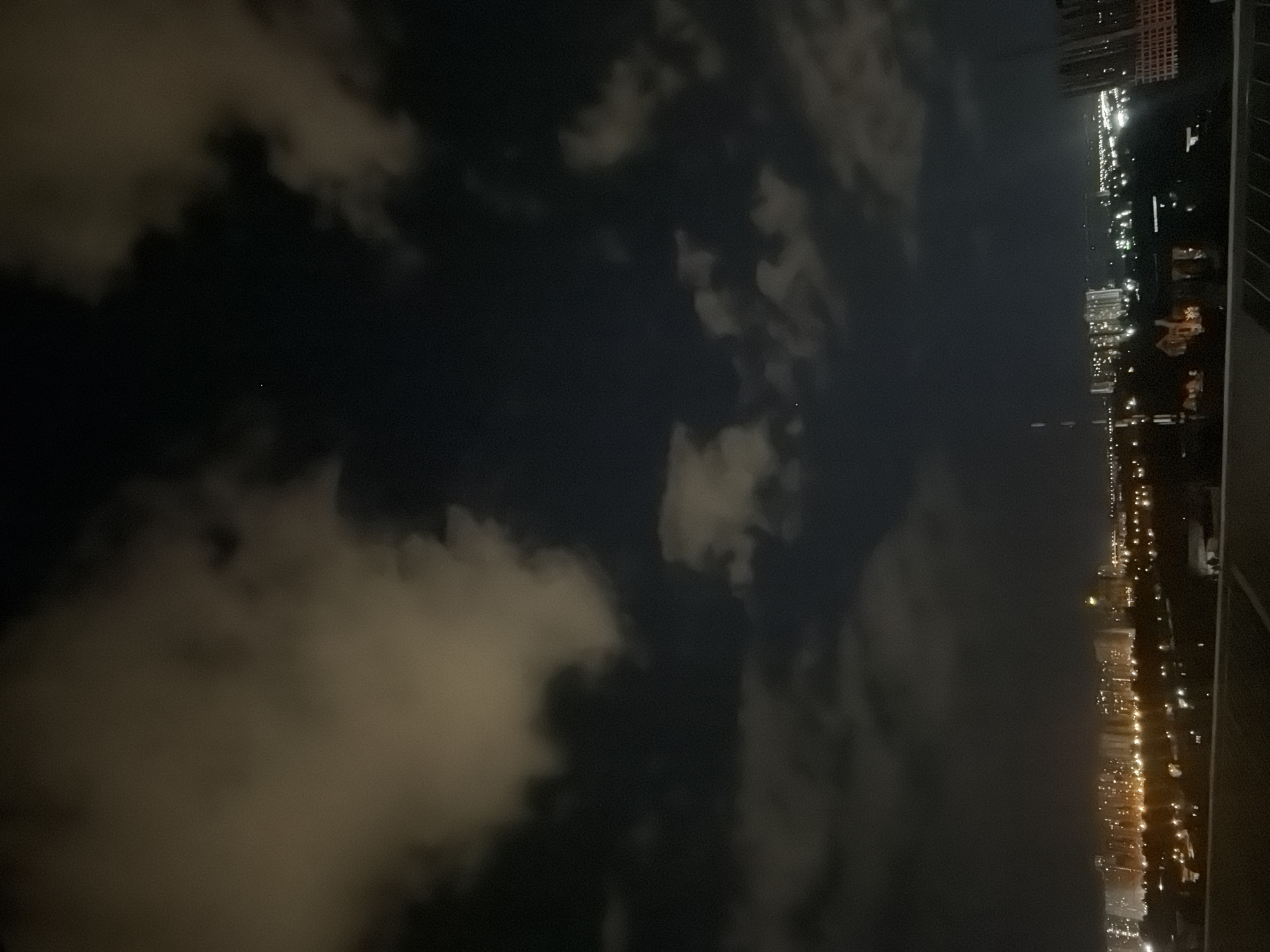 The image depicts a nighttime view of a cityscape, likely from an elevated position. The sky is dark with scattered clouds illuminated by ambient light. Below, a sprawling urban area is visible, characterized by numerous buildings with lights on, suggesting residential or commercial activity. The lights create a warm glow across the horizon. The architecture of the buildings is varied, with some appearing to be high-rise structures. The foreground shows a darker area, possibly a rooftop or balcony, from which the photograph was taken. There are no discernible people or specific actions captured in the image. The overall impression is of a quiet, nocturnal urban scene in a city setting. While the location is stated to be Kyiv, Ukraine, there are no specific landmarks or identifying features within the image to confirm this.