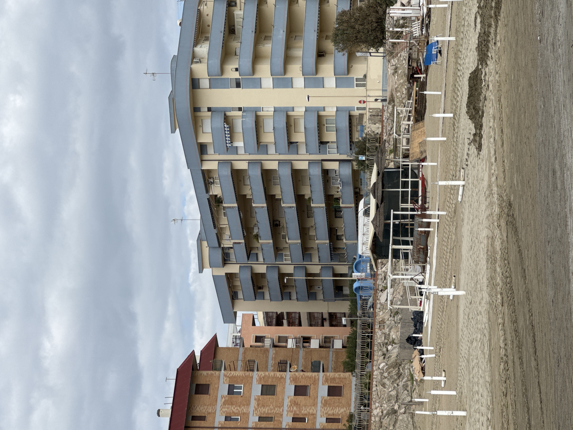 The image captures a coastal scene in Nettuno, Italy, featuring a row of multi-story buildings overlooking a sandy beach under an overcast sky.
The most prominent subject is a tall building with a light beige facade and distinctive blue-grey curved balconies, which create a wavy architectural pattern across its many floors. An antenna is visible on its roof. To the right of this building, other structures are present, including one with an orange-pink facade and another with a brick pattern and red-tiled roofs.
In the foreground, a sandy beach stretches across the frame. The beach appears to be in an off-season or transitional state, as beach furniture like umbrellas and chairs are largely disassembled or stored. Numerous white poles, likely bases for beach umbrellas, are stuck into the sand, and tire tracks are visible on its surface. A blue beach chair, some rocks, and what appears to be construction or storage materials are scattered along the edge of the beach. A metal fence separates the beach from the buildings.
The scene is static, with no human activity directly observed. The time of day is daytime, but the weather is cloudy and overcast, casting a soft, diffused light over the landscape.
Visible text includes a small blue sign with a white 'P' for parking on a pole near the fence, and a red circular sign nearby, though its text is unreadable.