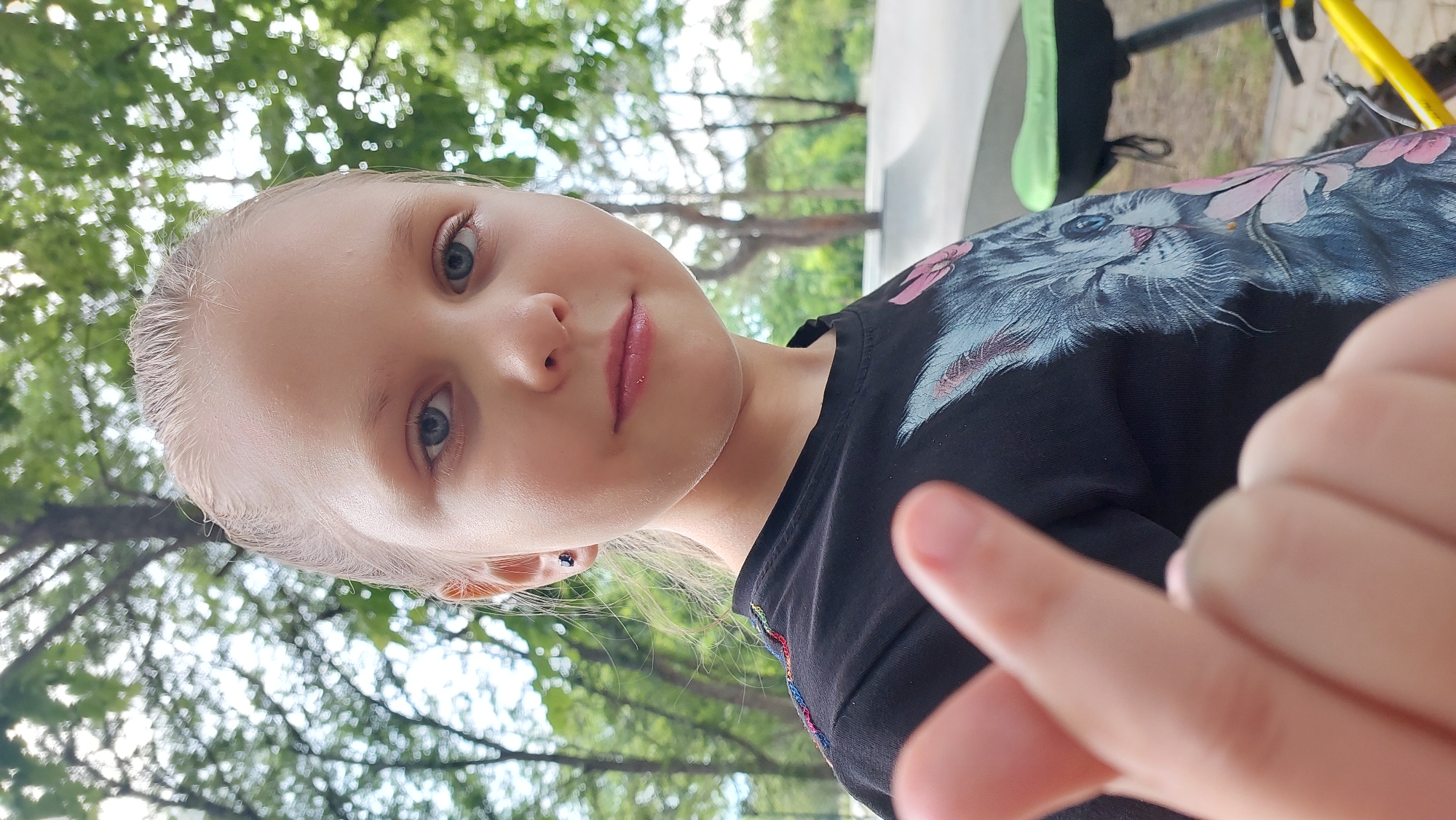 This is a close-up, low-angle, portrait-style photo of a young blonde girl. She is looking directly at the camera with a neutral expression. Her eyes are blue, and her lips are slightly parted. She is wearing a black t-shirt with a graphic of a cat with pink flowers on it. The background is blurred, but it appears to be outdoors with green trees and dappled sunlight. There are hints of a white structure and a yellow bicycle in the background. The image is slightly overexposed, and the lighting suggests it was taken during the daytime, possibly on a sunny day. The overall mood is casual and candid, as if it were a selfie. No visible text is present in the image.