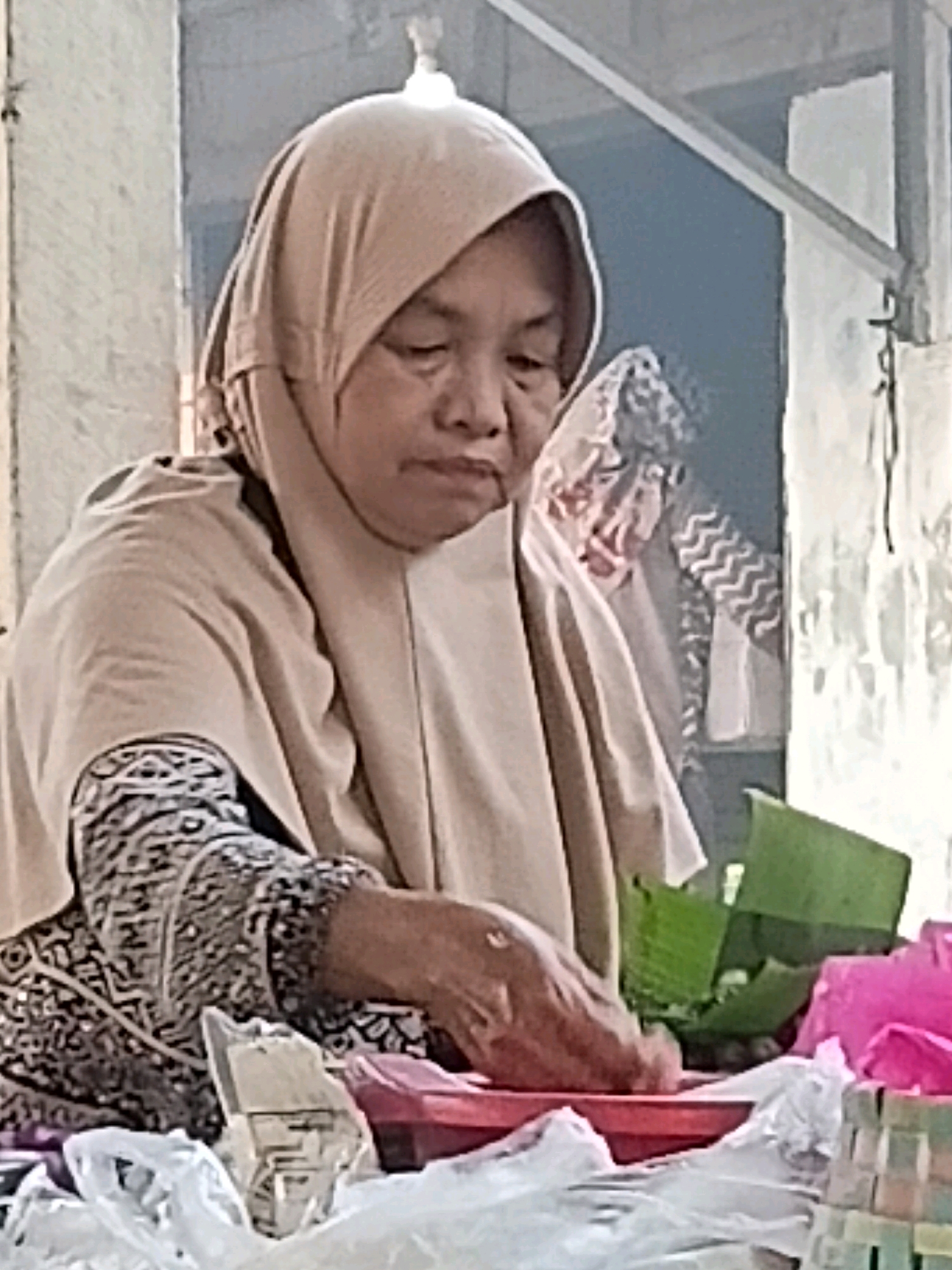 A woman wearing a beige hijab and a patterned black and white dress is seen from the chest up. She is looking down at her hands, which are engaged in preparing food. She is seated behind a red tray, where she is placing food wrapped in green banana leaves into small containers. To her left, there are plastic bags and a white object that appears to be a food container. To her right, there are pink items and a woven basket. The background is blurred, but suggests an outdoor market stall in Margo Rukun, Indonesia.