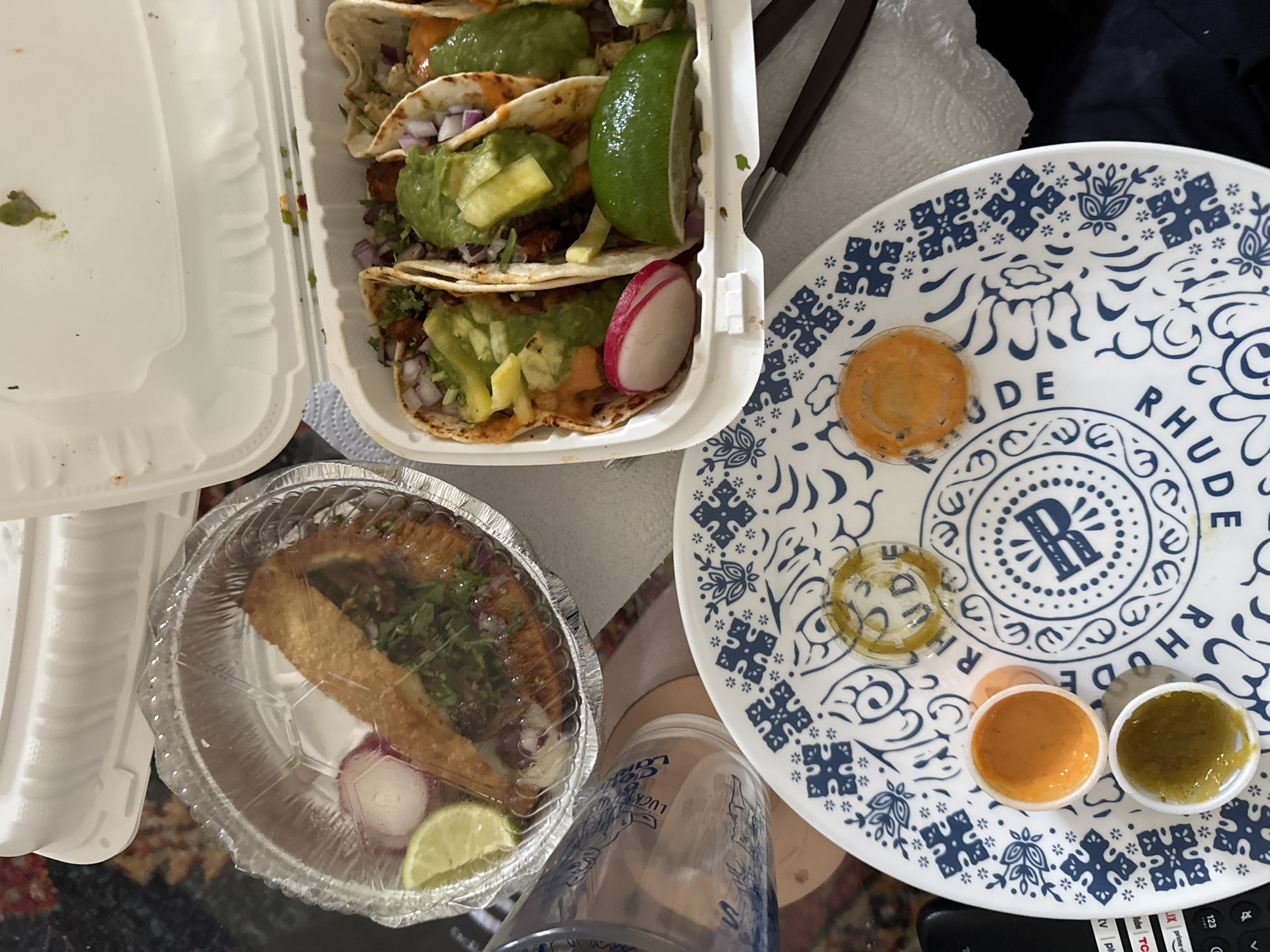 The image shows an arrangement of takeout food containers on a table. A white clamshell container holds several tacos filled with meat, onions, cilantro, avocado, and a lime wedge. Next to it is a circular foil container holding what appears to be a fried quesadilla with toppings and a lime wedge. A plate with blue and white floral patterns sits to the right, holding small containers of different sauces. A clear plastic cup with a graphic is also present. Based on the name "Rhude" on the plate, this may be a scene related to a business, but I cannot confirm a location in the city of New York.