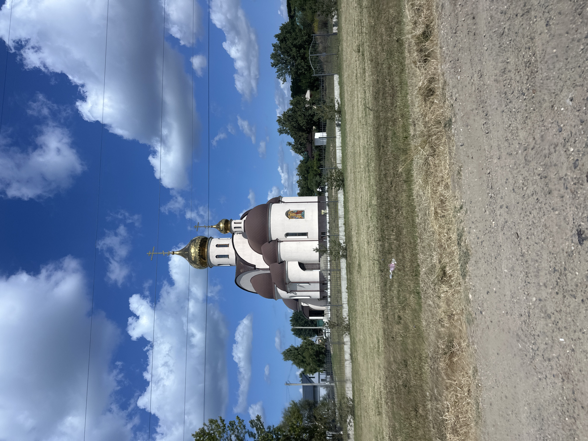 A view of an Orthodox church, with white walls and brown and gold domes, in Sennoy, Russia, under a bright blue sky with scattered white clouds. The church is situated behind a fence and a field of dry grass. Power lines run horizontally across the top of the image. In the foreground, a dirt road is visible. The overall scene depicts a rural or suburban setting on a sunny day.