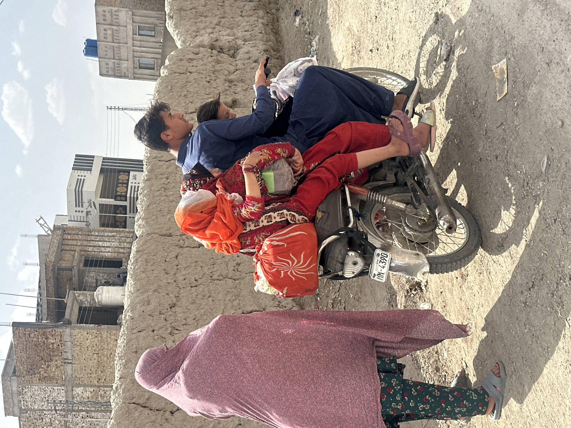 In Quetta City Tehsil, Pakistan, a family of three is crowded onto a motorbike. The man, wearing a blue shirt, is sitting in the back looking at a phone. A woman is in front of him wearing a red, printed dress and orange headscarf. A third person is in front of her. Next to the motorbike a person, partially covered in a pink shawl, is wearing a floral-patterned outfit and grey slippers. The motorbike has a license plate that reads "MM-4390." The setting appears to be a roadside with unfinished buildings in the background. The ground is a mix of dirt and concrete.