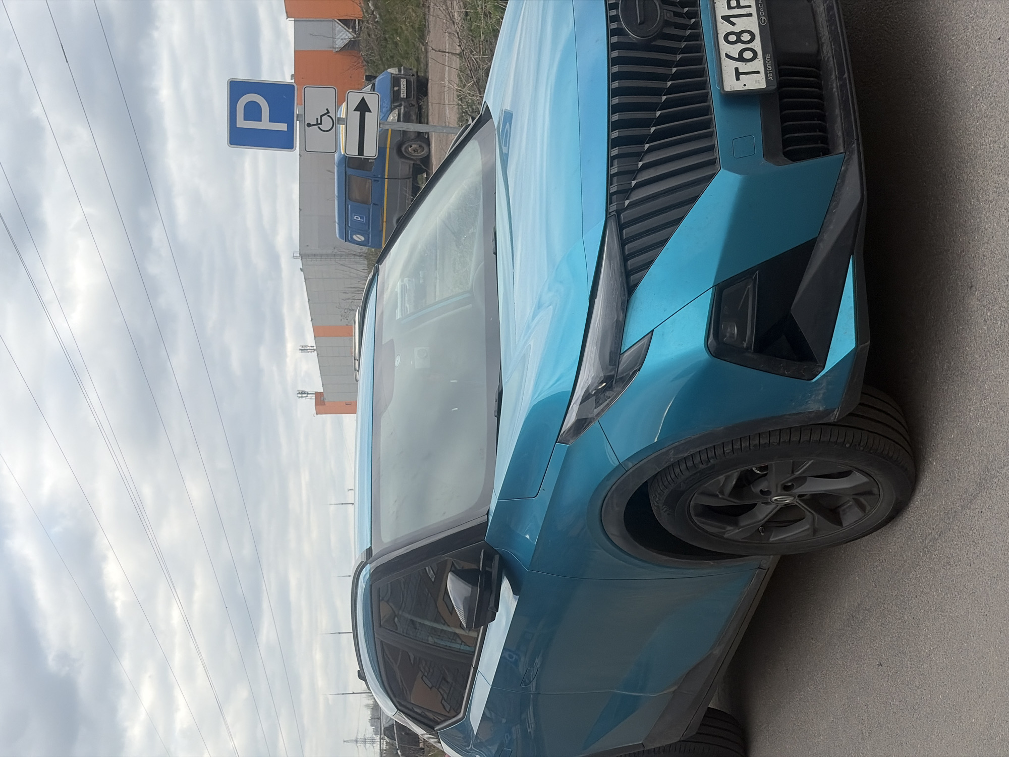 The image displays a teal-blue SUV parked on an asphalt surface under an overcast sky, indicating daytime. The vehicle is captured from a front-left perspective, highlighting its modern design with a prominent dark grille and angular headlights. A Russian license plate, visible as "T681P" with "АВТОТОТЕ" (AUTOTOTE) printed on its frame, is affixed to the front. The SUV features dark, multi-spoke wheels.
In the background, positioned to the left of the SUV, a cluster of road signs is visible. These include a blue square "P" (parking) sign, a white sign with a black disabled parking symbol, and a white rectangular sign with an upward arrow, likely indicating accessible parking. Further back, a blue van or bus-like vehicle, partially showing a Russian license plate "H394PK" and a "P" symbol on its side, is parked near some sparse greenery. Beyond the vehicles, industrial or commercial buildings, one with an orange facade, are discernible. Power lines crisscross the cloudy sky. The scene is static, depicting a parked vehicle in what appears to be an urban or suburban setting in Saint Petersburg, Russia. No individuals are present, and no particular activity or emotional state is conveyed.