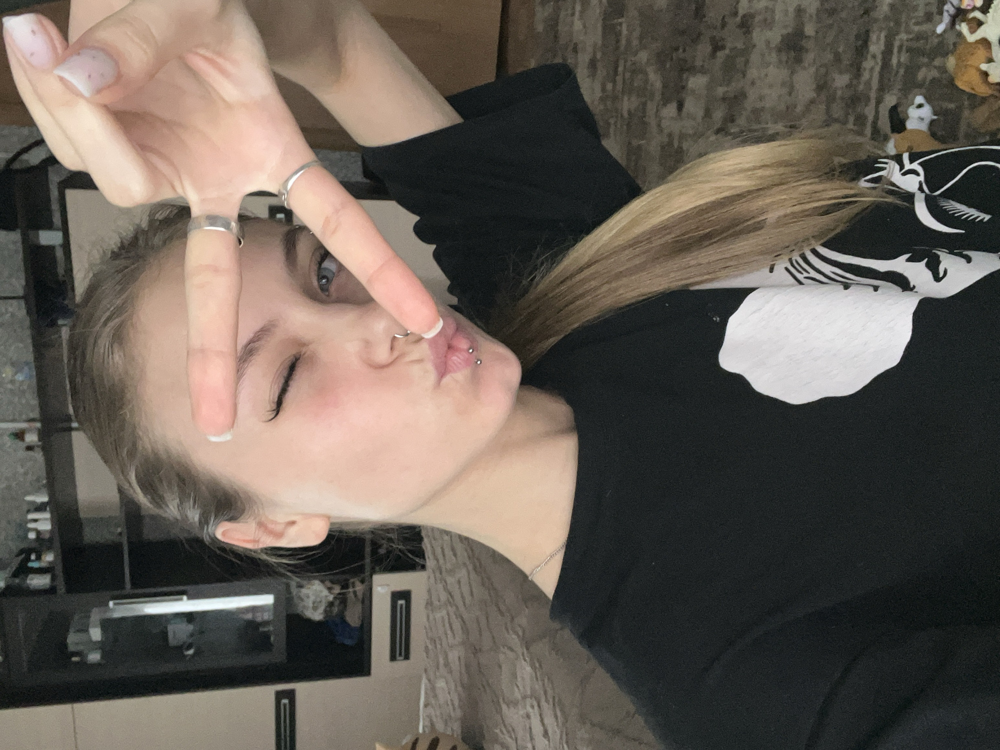 A young woman poses indoors, making a peace sign with her fingers. She winks her left eye and puckers her lips, wearing rings on two fingers. She has a nose piercing and lip piercings. Her blond hair is pulled back. She is wearing a black long-sleeved shirt with a white design. A kitchen or storage area is visible in the background, with a beige wall and dark cabinets. A textured wall is partially visible. The image suggests a casual selfie taken indoors. The location, Фоки, Russia, is irrelevant as there is no visual information to connect the image to the city.