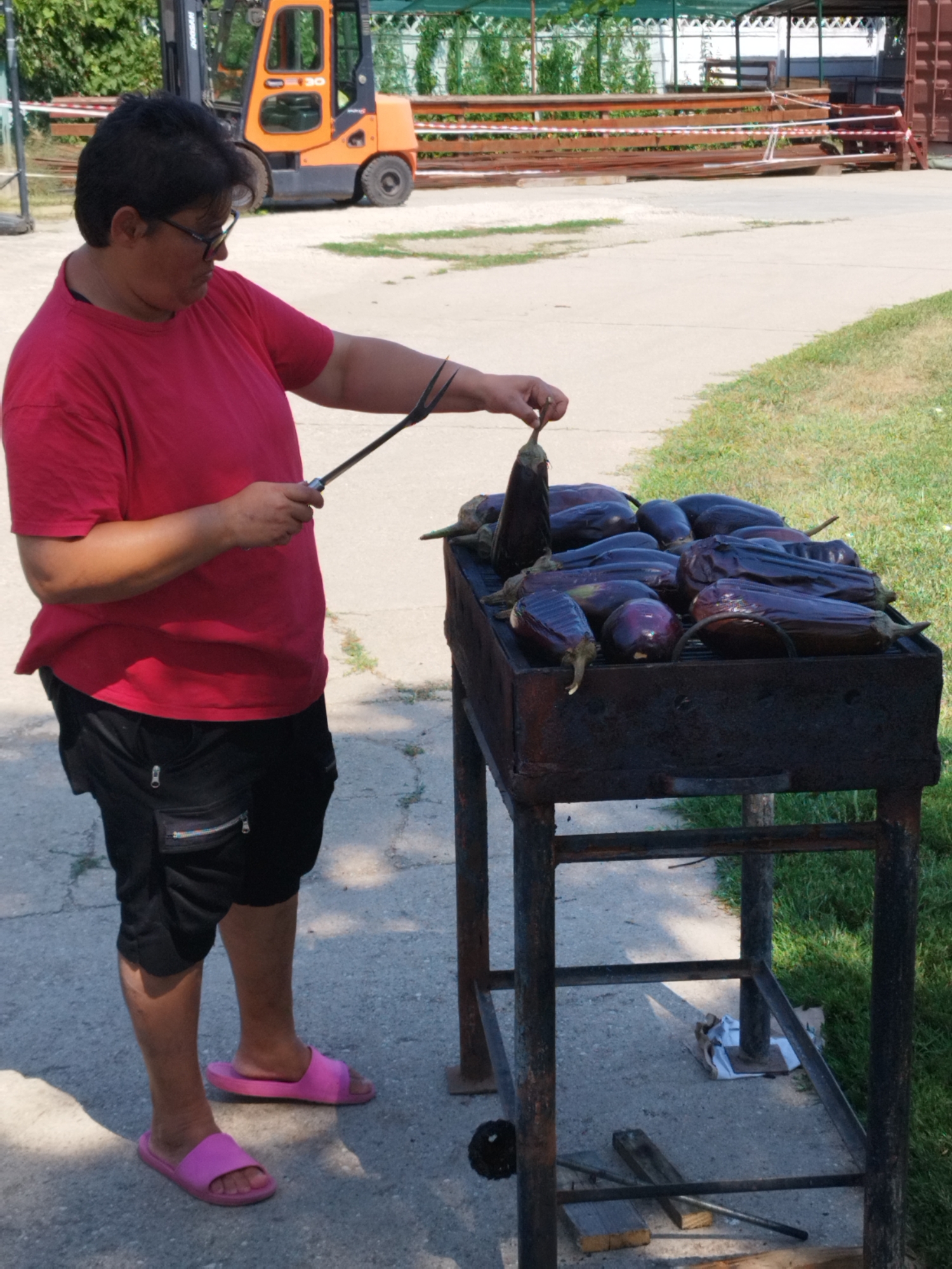 A woman wearing a red t-shirt, black cargo shorts, and pink slides is preparing to grill a batch of eggplants. She is holding one eggplant with a fork, poised to place it on a dark metal grill filled with more eggplants. The grill is situated on a concrete surface, with green grass visible to the right. In the background, an orange forklift is parked, suggesting an outdoor industrial or storage area. The scene appears to be in Bucharest, Romania, possibly a food preparation area or an outdoor event.