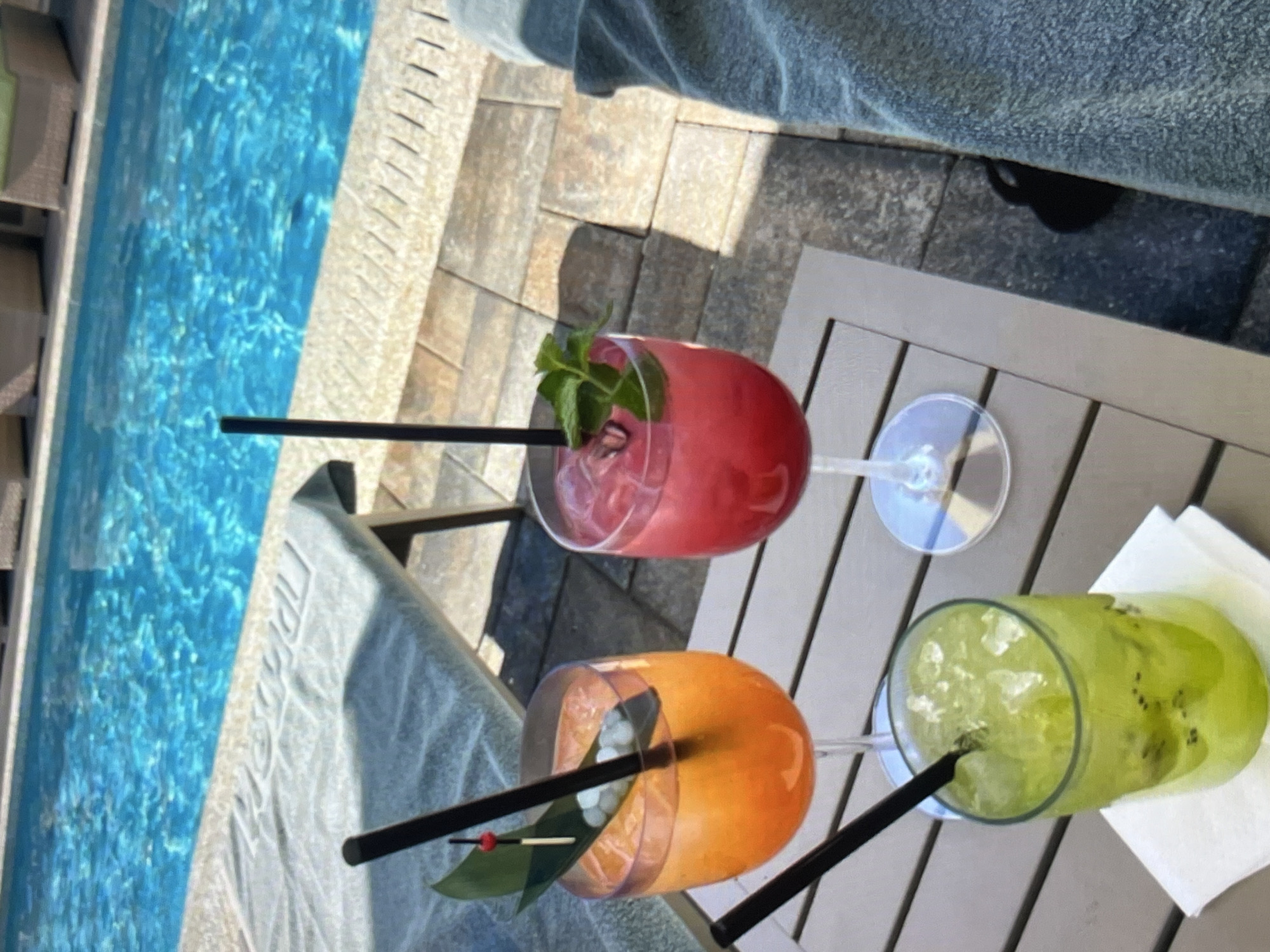 This media file features a close-up view of three colorful cocktails resting on a wooden table next to a swimming pool. The scene is set outdoors, with the bright blue water of the pool occupying the left side of the frame. The table has a slatted surface, and the ground around it appears to be paved with stone tiles.
The cocktails are presented in different types of glassware. The first, closest to the viewer, is an orange-colored drink served in a wide-mouthed glass, garnished with a leaf and a small red berry on a skewer. A black straw extends from this drink. To its right, a green cocktail is served in a more traditional glass, filled with ice and visible slices of kiwi fruit. This drink also has a black straw. Further back and slightly to the left, a third cocktail, a vibrant pink or red, is in a stemmed glass. It's garnished with a sprig of mint and also contains a black straw.
The lighting suggests it is daytime, likely sunny given the shadows cast on the ground. The overall mood is relaxed and suggestive of a vacation or leisure activity, possibly a poolside lounge. There is a hint of a folded towel in the upper part of the image, adding to the atmosphere of relaxation. No people are visible, but the presence of the drinks and the pool strongly implies their recent or imminent activity. There is no visible text in the media file.