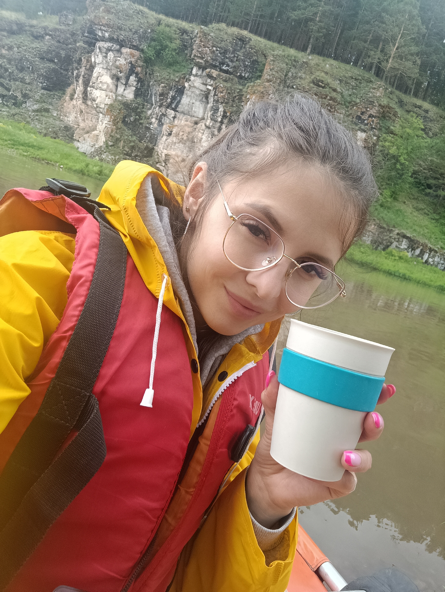 A woman is taking a selfie in a canoe on the Yuryuzan River near Idrisovo, Russia. She is wearing a yellow waterproof jacket over a red and orange life vest. She has glasses on and is holding a white travel mug with a teal band. The background shows a rocky cliff face and a forested hillside. The river is calm and murky.