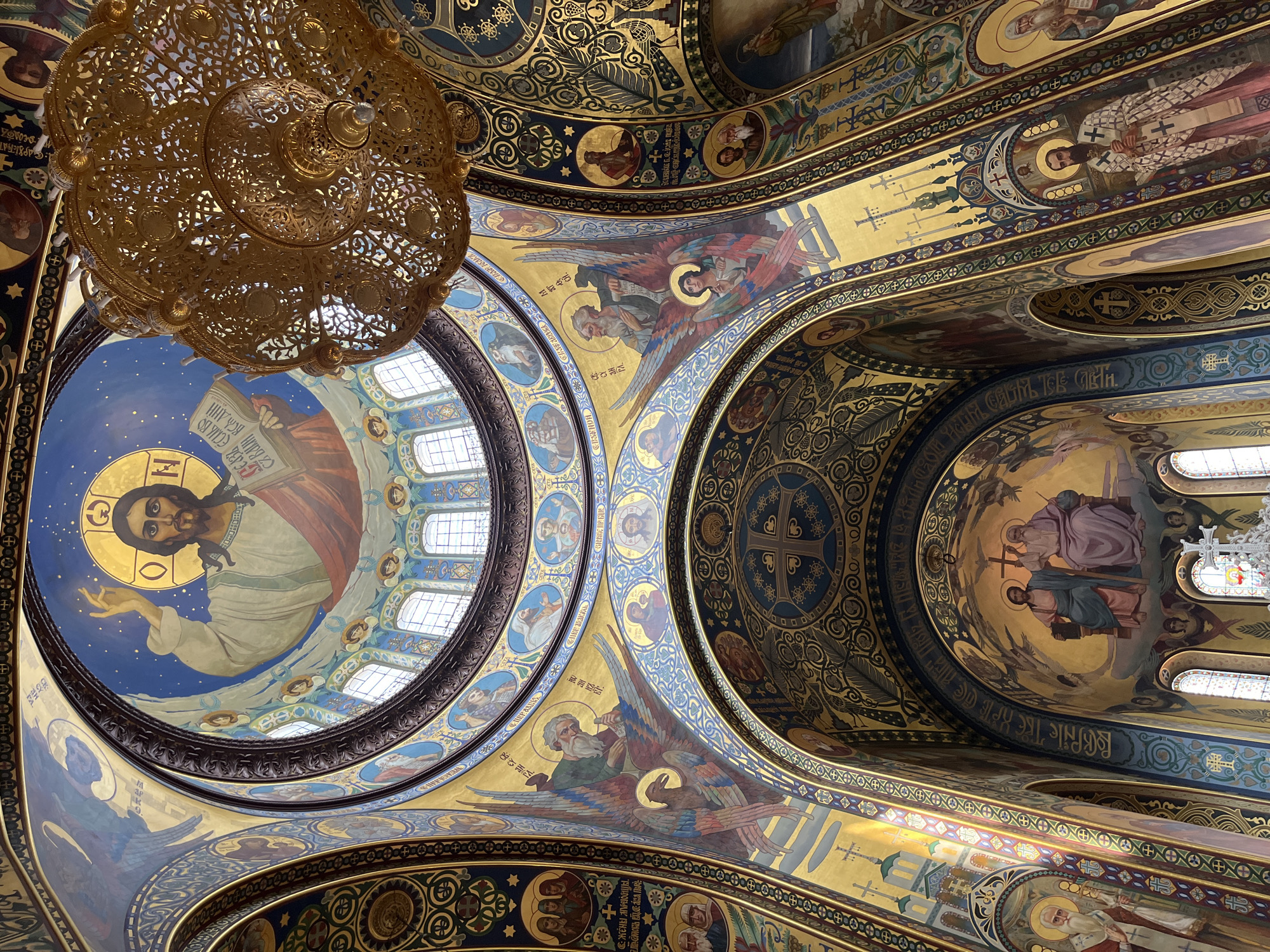 The image shows the interior of the Ascension Cathedral in Novocherkassk, Russia. The cathedral is ornately decorated with religious frescoes covering its domed ceiling and walls. A large, golden chandelier hangs from the ceiling. The frescoes depict scenes from religious narratives, including figures of Jesus Christ, angels, and saints. The architectural style is characteristic of Russian Orthodox churches, with elaborate gold accents and vibrant colors. The lighting suggests it is daytime, with light entering through arched windows.