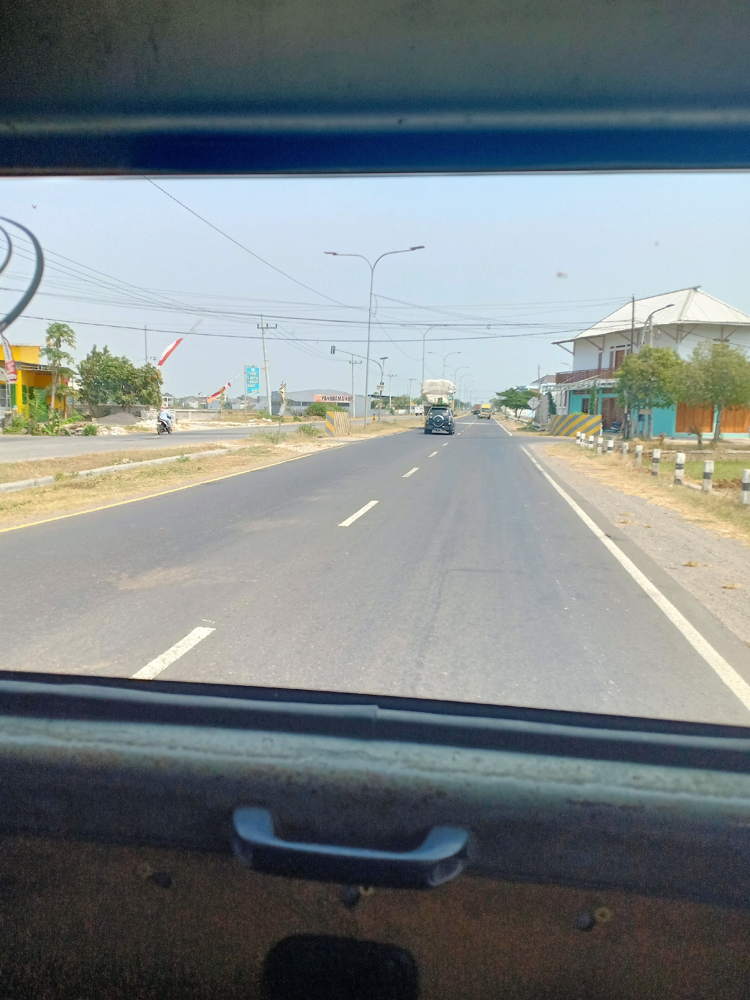 The image is a point-of-view shot from inside a vehicle looking out at a road in Turigede, Indonesia. A two-lane asphalt road stretches into the distance, with white dashed lines marking the lanes. The sky is a clear, pale blue. On the right side of the road, a white building with a prominent white roof and an orange trim is visible, along with trees and a line of white posts with black tops. Further down the road on the right, a yellow and blue striped barrier is present. On the left side of the road, there are various buildings, including a yellow and green structure. A motorcycle with a rider is seen on the left side of the road. A black SUV with a trailer is driving in the distance on the left lane, and a yellow truck is further ahead. Overhead power lines are visible across the scene. The foreground shows the interior of the vehicle, with a dark, textured surface and a dark gray handle.