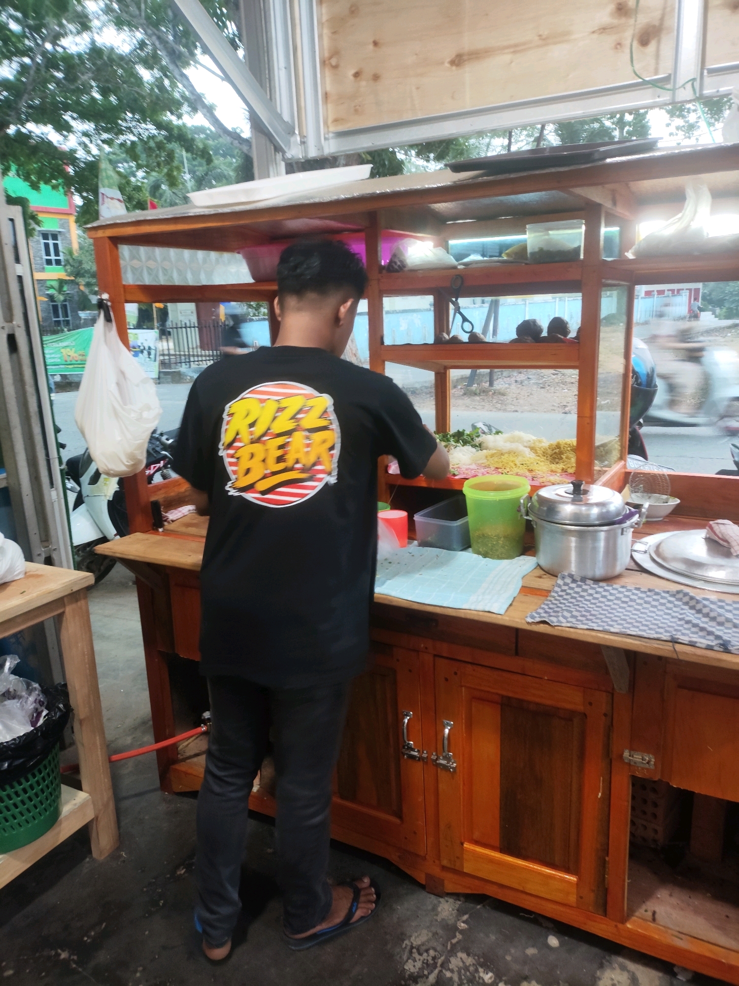 The image depicts a food stall in Palembang, Indonesia, likely on a street or roadside. A person, seen from behind, is wearing a black t-shirt with a yellow and red circular graphic that says "RIZZ BEAR". They are standing behind a wooden food cart, which is laden with various ingredients, including what appears to be noodles, vegetables, and condiments. A large stainless steel pot is prominently displayed on the counter. The stall has a transparent front, allowing a view of the ingredients. In the background, there are trees, a building with green and red accents, and traffic, including a motorcycle. The overall scene suggests a typical street food vendor operating in a busy urban environment.