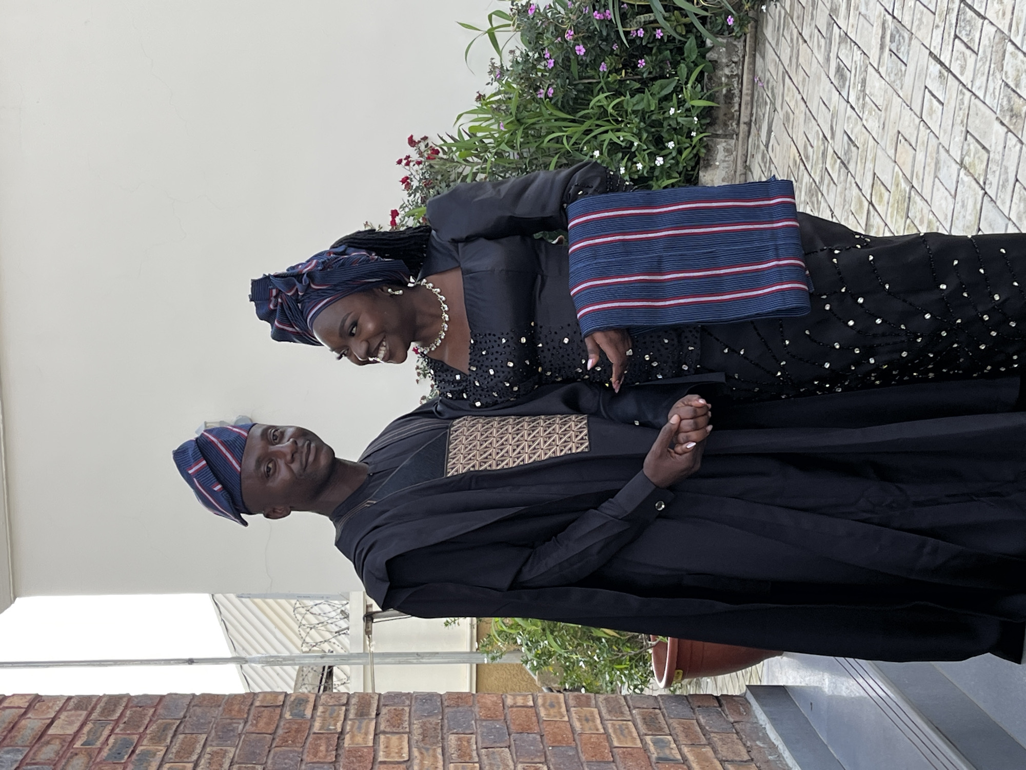 A Black couple dressed in traditional Nigerian attire poses for a photograph outdoors. The man, on the left, wears a black agbada, a wide-sleeved robe, over a black shirt with a geometric woven pattern on the chest. He also sports a matching fila, a cap, with navy blue, red, and white stripes. The woman, on the right, is adorned in a black dress with puff sleeves and intricate beading. She wears a matching headwrap, similar in color and pattern to the man's cap, and a diamond necklace. They are holding hands and smiling at each other. Behind them, a white wall and lush green plants with small red and pink flowers create a natural backdrop. The ground is paved with light-colored bricks. The couple is positioned in front of a brick building. The scene appears to be in Gura Top, Nigeria.