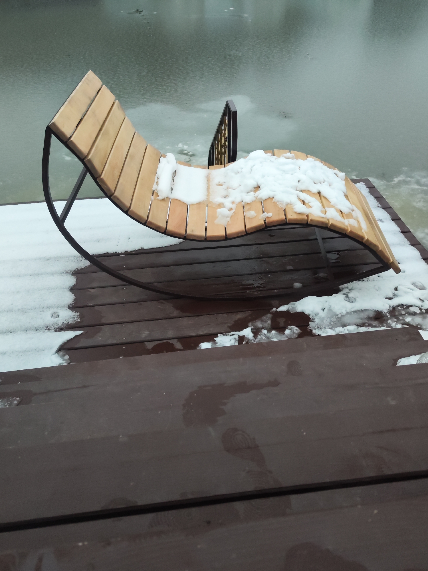 The image depicts a partially snow-covered wooden rocking chaise lounge on a wooden deck. The deck appears to be adjacent to a body of water, possibly a river or lake, with some ice visible on the surface. The location is Chaikovsky, Russia. There are no people visible in the scene. The focus is on the furniture and its wintry setting. The deck is elevated, with steps leading down to it. A small portion of a wrought iron railing is visible behind the chaise.
