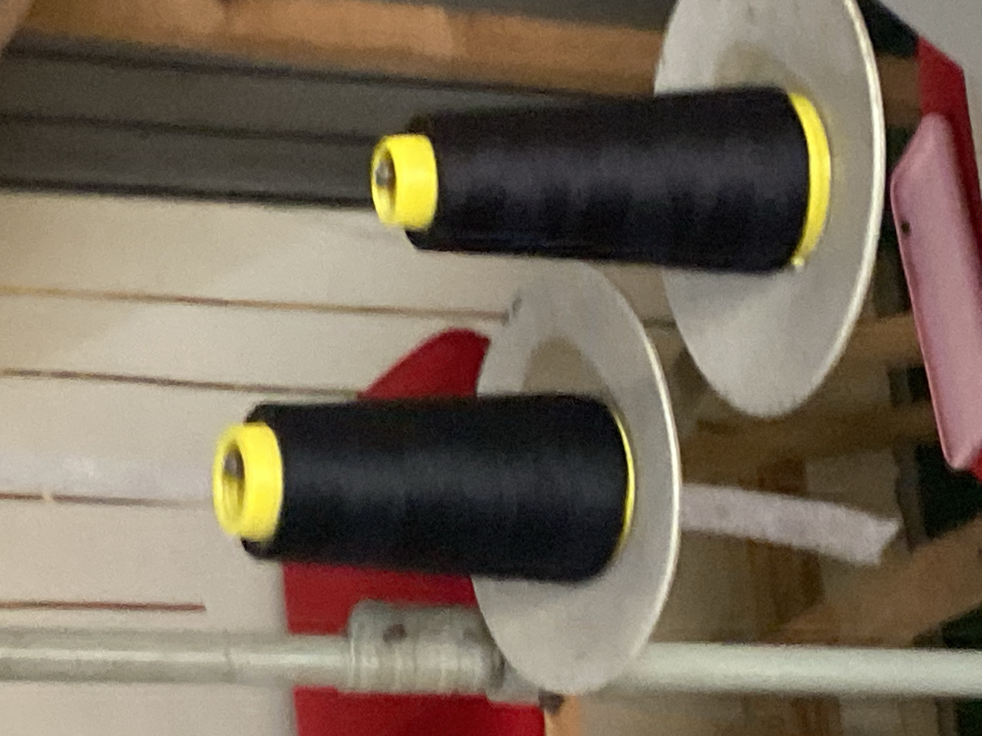 The image shows two large spools of black thread with yellow caps, placed on circular metal stands. The spools are conical in shape and are the primary focus of the image. The background is blurred, but appears to be an indoor setting with vertical lines suggestive of shelving or a wall. There are also hints of red fabric in the background. A pink object, possibly a phone case, is visible in the foreground. The image does not provide any contextual information about its location in Jauro Yinu, Nigeria, or any other specific geographical details.