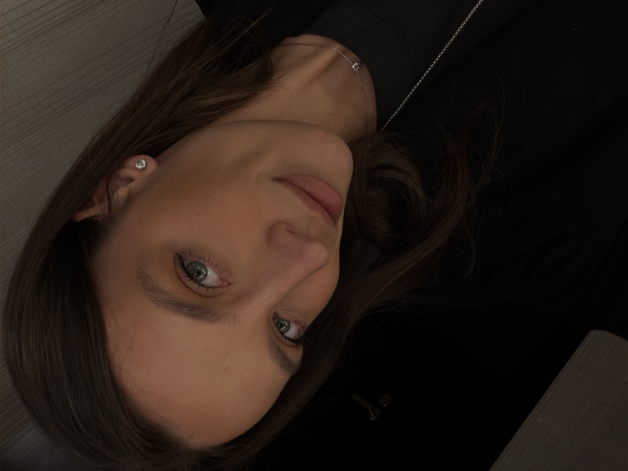 A close-up, slightly tilted, low-lit photograph depicts a light-skinned woman with brown hair. She has light-colored eyes and is wearing a small stud earring and a delicate silver necklace. The background is predominantly dark, with hints of structured surfaces in light gray and black fabrics or materials. The image does not provide information about the location in Budva, Montenegro.