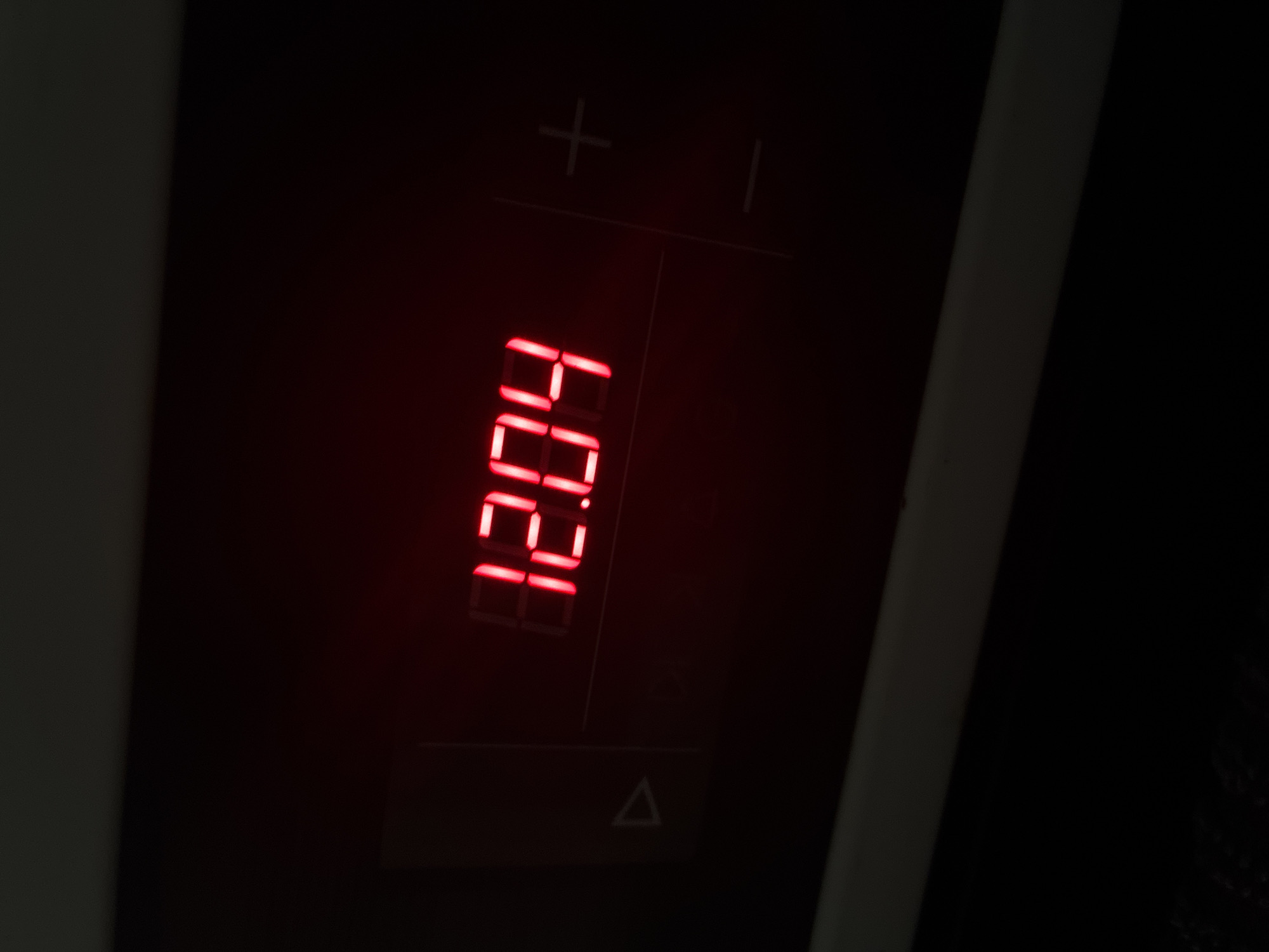 An extreme close-up shot shows a digital clock displaying the time 12:04 in red LED numerals. To the left of the clock, a triangle icon is visible, and to the right, plus and minus symbols are faintly etched into the dark surface. The scene is dimly lit, with the red glow of the clock being the primary light source.