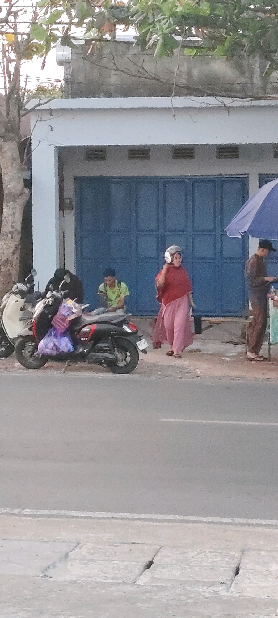 In Bentok Kampung, Indonesia, a street scene unfolds with a building featuring blue folding doors and a grey concrete wall above. A motorcycle, white and black, is parked on the side of the road, with a purple bag strapped to its rear. To the right of the motorcycle, a young man in a green shirt is seated, looking at a device. Further right, a woman in a pink headscarf and a long pink dress stands, holding a white helmet to her ear. A man in a brown shirt and pants stands near a stall under a blue umbrella, with food items visible on a surface. The ground is a mixture of dirt and pavement, and the road in the foreground shows white lane markings. Trees with green and brown leaves overhang the scene.