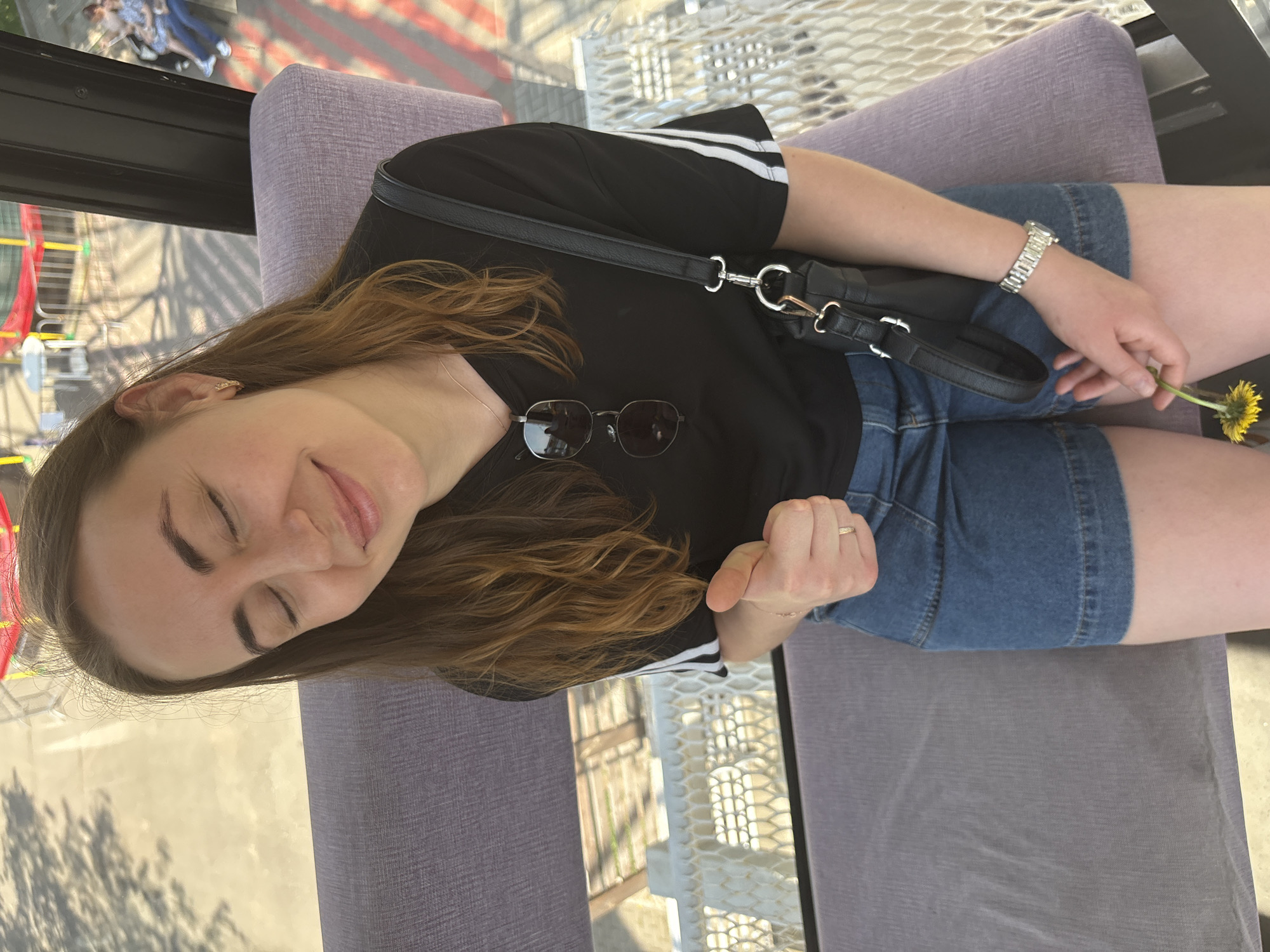 A young woman is seated, holding a yellow flower, in a booth on the Izhevsk Ferris Wheel in Izhevsk, Russia. She has brown hair and is wearing a black t-shirt with white stripes on the sleeves and blue denim shorts. She has a black purse slung over her shoulder and a silver watch on her wrist. A pair of sunglasses rests on her shirt. The background reveals the city from the Ferris wheel's elevated perspective.