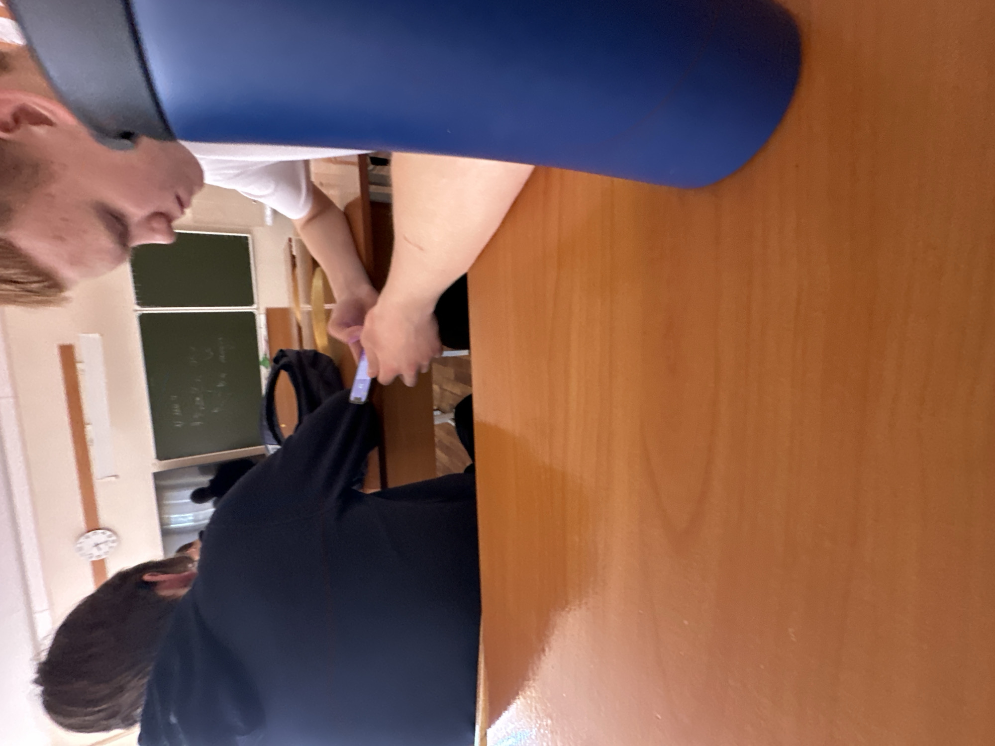 A classroom scene shows two individuals seated at a wooden desk. One person is visible from the shoulders up and wears a dark blue or black sweater, with dark hair. The second person is only partly visible, wearing a white shirt with a partial face visible. The scene is slightly obscured by a blue object hanging overhead. In the background, there's a green chalkboard and a clock on the wall. The exact location and city are not identifiable from the image.