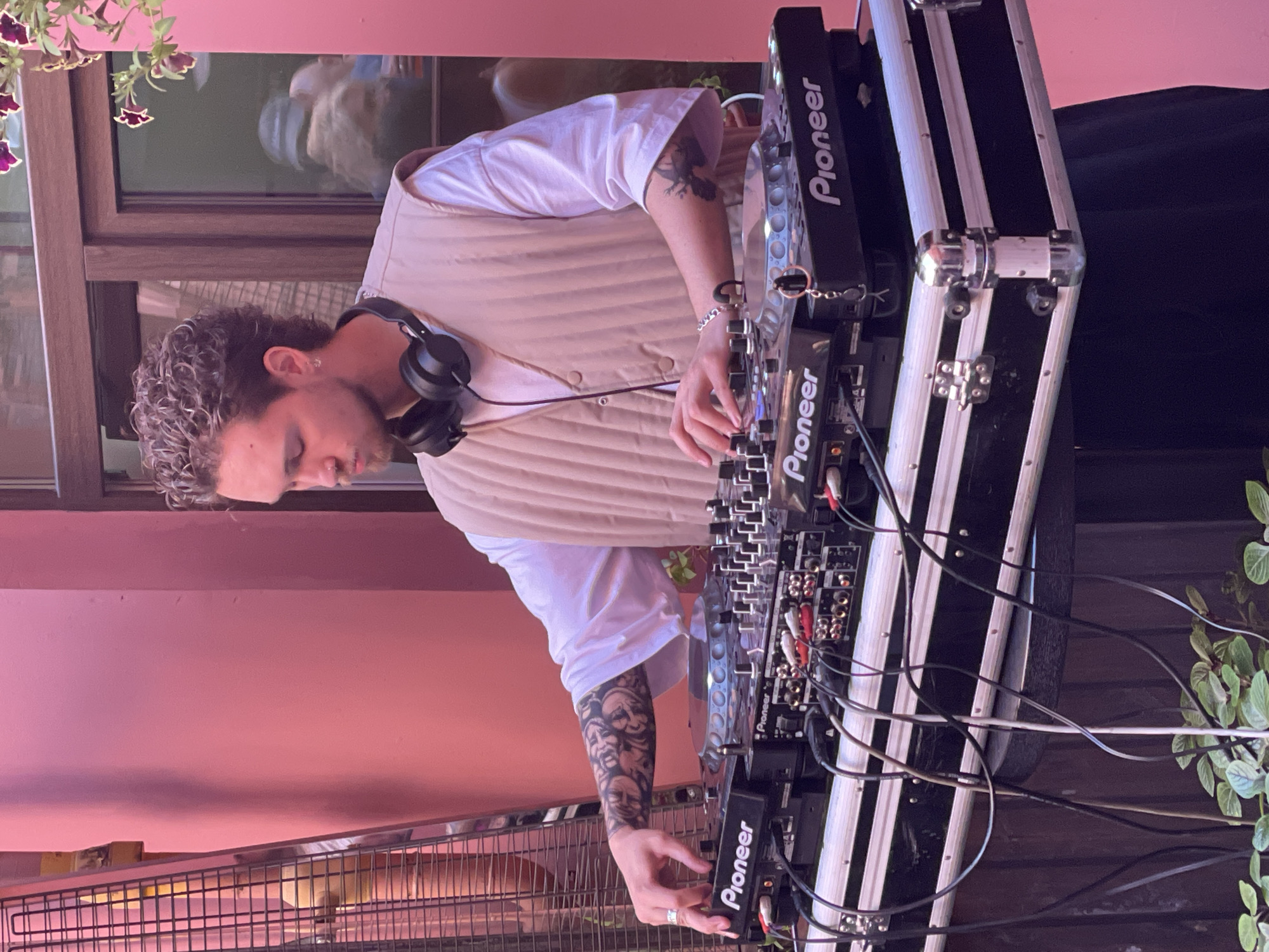 In Saint Petersburg, Russia, a young man with curly brown hair and tattoos on his arms is operating a DJ setup. He is wearing headphones around his neck and a beige vest over a white shirt. His left hand is touching a dial on the DJ equipment, which is placed in a black case with silver accents. The background features a pink wall and a window reflecting other people. Green plants are visible at the bottom of the image.
