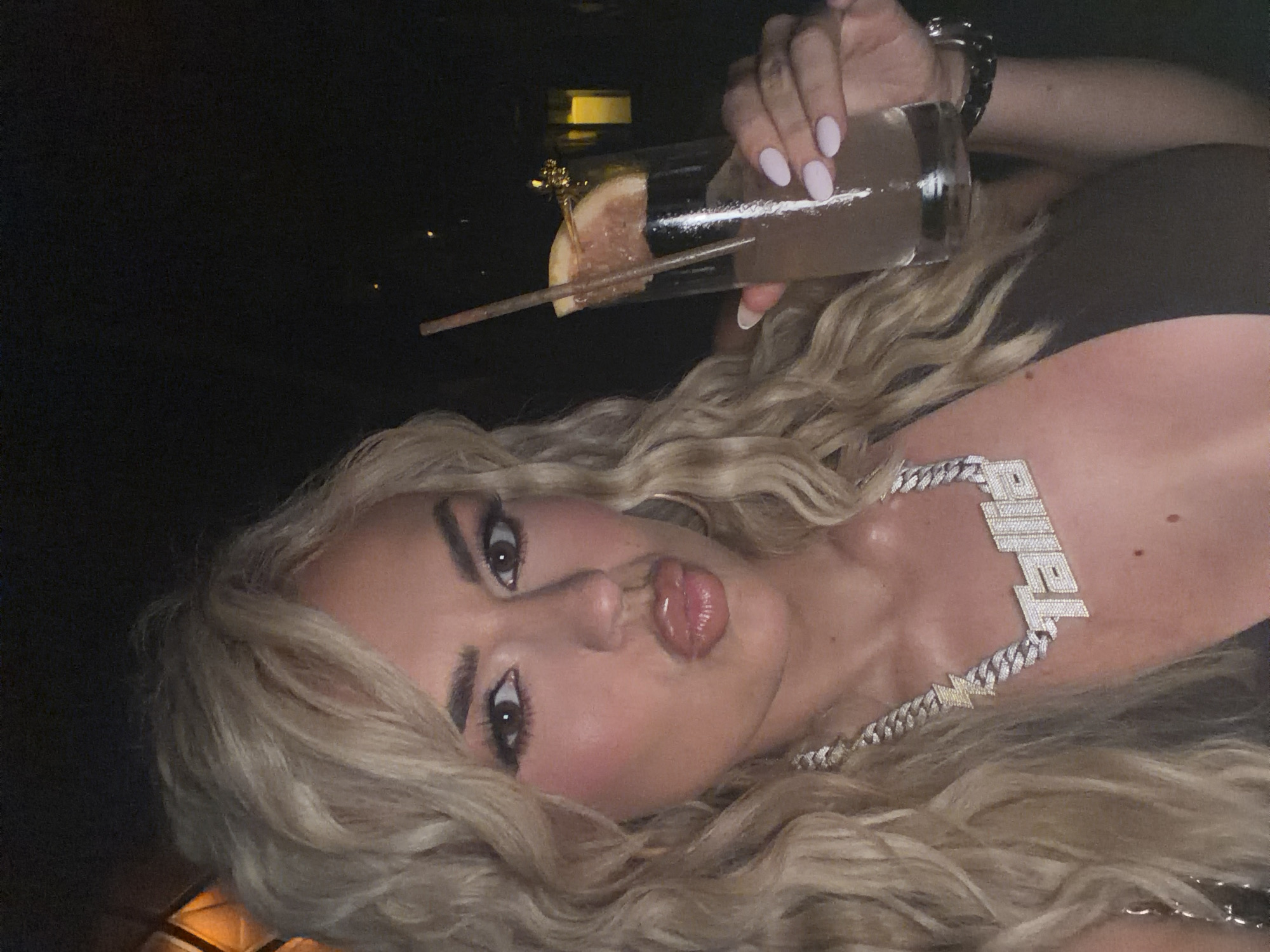 A young woman with blonde, wavy hair lies on her side and looks at the camera. She is wearing a large diamond chain necklace that spells out the word "Tahila" and has full lips. In the upper portion of the picture, a hand holds a tall, clear glass filled with a light-colored liquid and garnished with a slice of citrus, pierced by a straw and a small, decorative pick. The background appears dark, though a slight glow suggests a possible light source behind the glass. No specific location is identified, though the prompt specifies Miami Beach, United States of America.