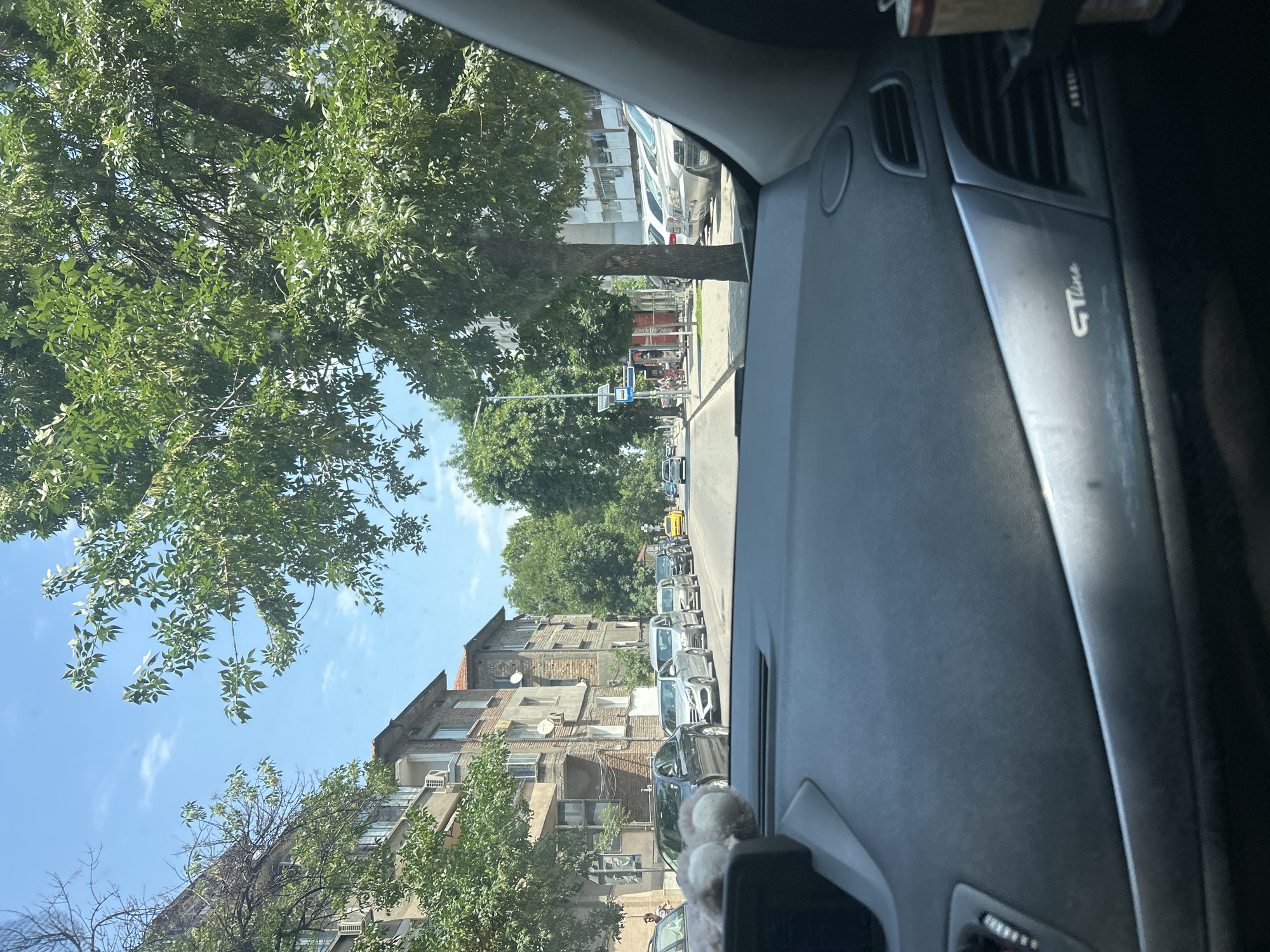 The image shows a view from the interior of a car driving down a city street in Plovdiv, Bulgaria. The street is lined with buildings and trees. The sky is blue with some clouds. In the foreground, the dashboard of the car is visible, including an air vent and a "GT Line" badge. In the middle ground, there are several cars parked along the side of the road and a line of cars in traffic. In the background, there are more buildings and trees, and a yellow taxi can be seen in the distance. There are a few pedestrians visible on the sidewalk.