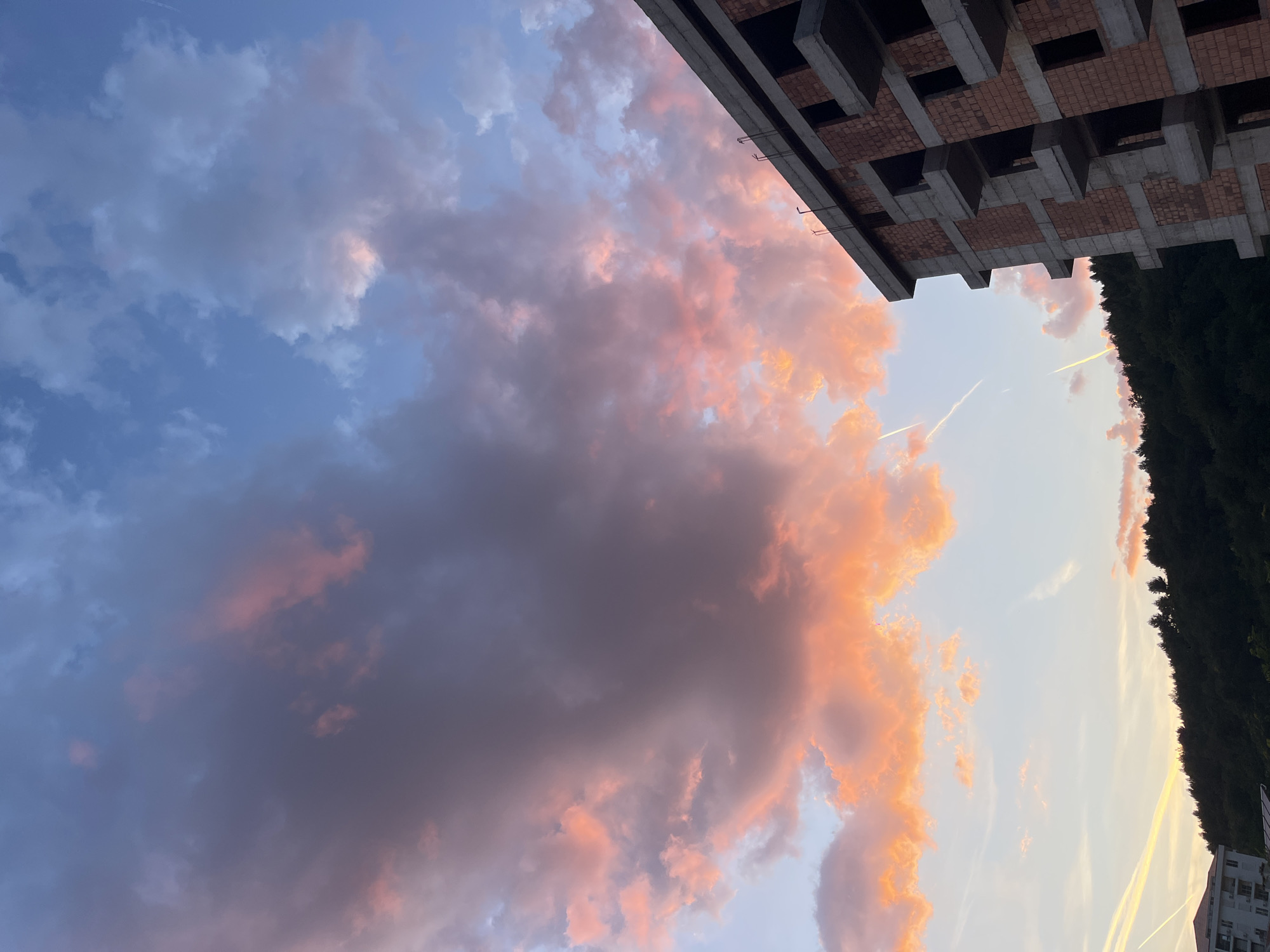 The image shows a sky at dusk or dawn in Rafailovići, Montenegro. Pink and orange clouds fill the center and right of the frame, transitioning into a blue sky on the left. An unfinished brick and concrete building structure occupies the top right corner, partially obscuring the sky. A line of trees lines the edge of the right side of the frame, near another building. Two contrails are visible in the sky.