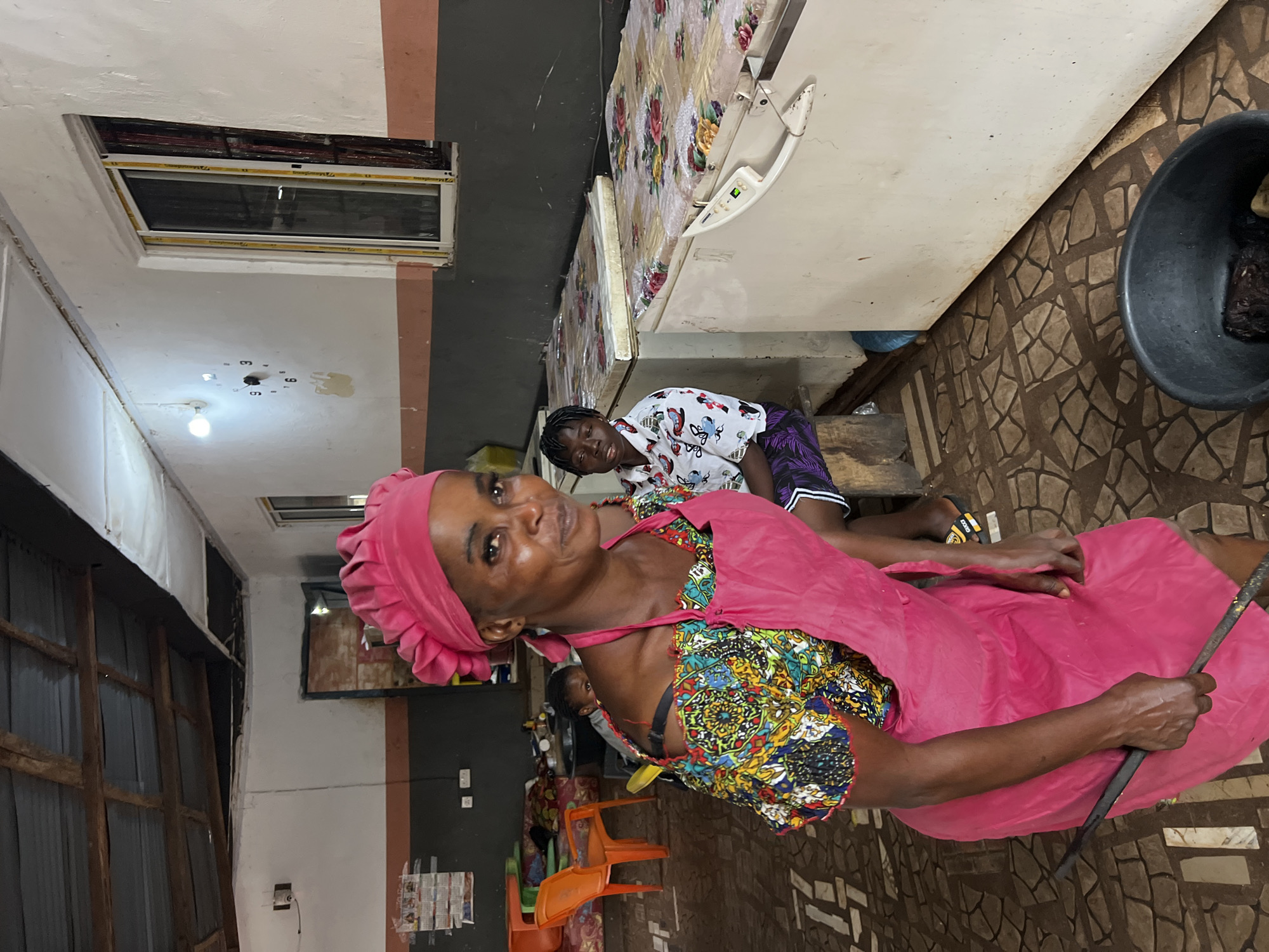 The image depicts an indoor scene, likely a food stall or a small eatery, in Nkpor, Nigeria. In the foreground, a woman wearing a vibrant, patterned off-the-shoulder dress and a pink apron and headscarf is looking towards the camera. She is holding a long, dark metal tool in her right hand. Behind her, another young woman is seated, wearing a white shirt with a cartoon-like print and dark pants. The background reveals industrial-looking freezers covered with floral patterned cloths, suggesting a commercial operation. The room has a corrugated metal ceiling and concrete walls painted with distinct horizontal bands of color. The floor is tiled with a mosaic pattern. In the distance, orange plastic chairs are visible, and the general ambiance suggests a bustling, everyday commercial environment.
